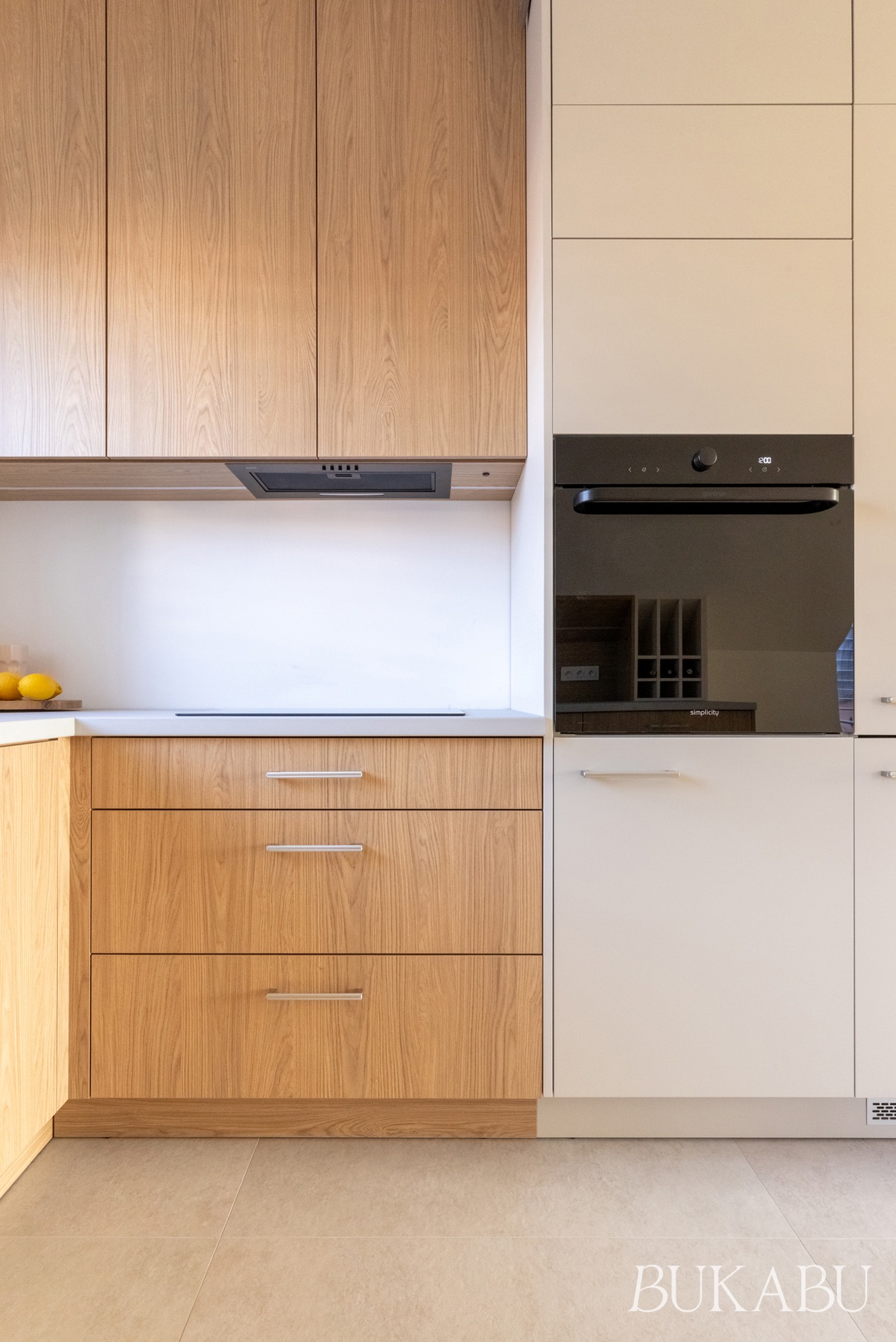 Jasna kuchnia z drewnianymi frontami szafek i białymi elementami. Widoczny piekarnik w zabudowie. Minimalistyczny design, naturalne światło. Ujęcie na detale zabudowy kuchennej.