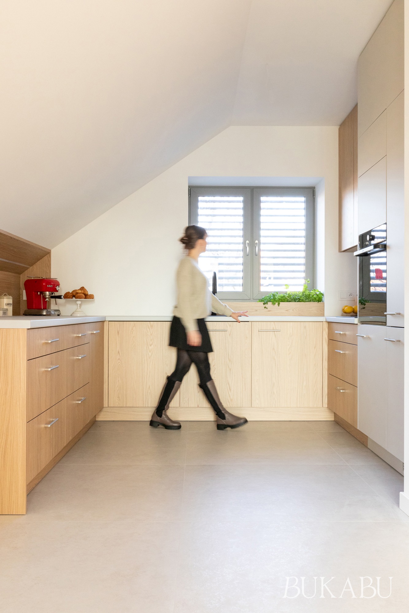 Jasna kuchnia na poddaszu z drewnianymi frontami szafek, szarym oknem z żaluzjami i czerwoną maszynką do kawy. Kobieta przechodzi przez kadr, dodając dynamiki.