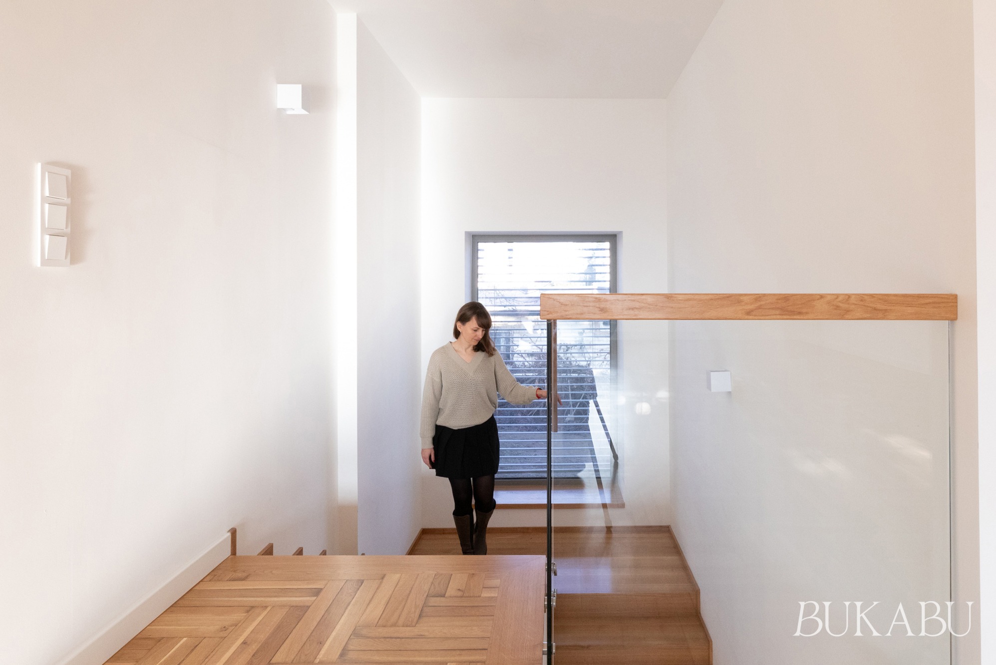 Jasne wnętrze domu po renowacji: drewniane schody z parkietem w jodełkę, szklana balustrada z drewnianym elementem, kobieta schodząca po schodach, minimalistyczne oświetlenie.