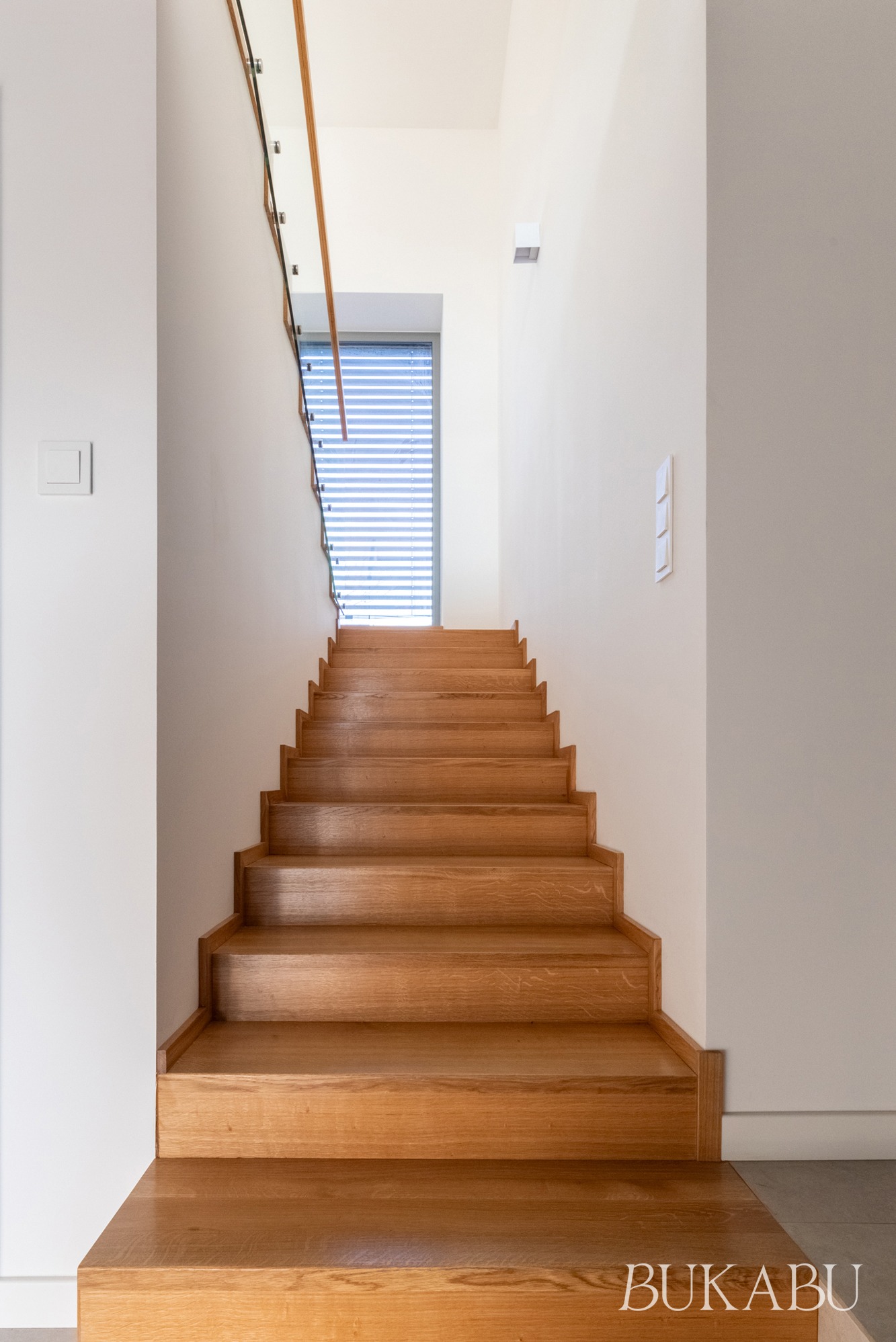 Drewniane schody z przeszkloną balustradą w jasnym wnętrzu. Ujęcie od dołu, widoczne okno z żaluzjami w tle. Minimalistyczny design, naturalne światło.