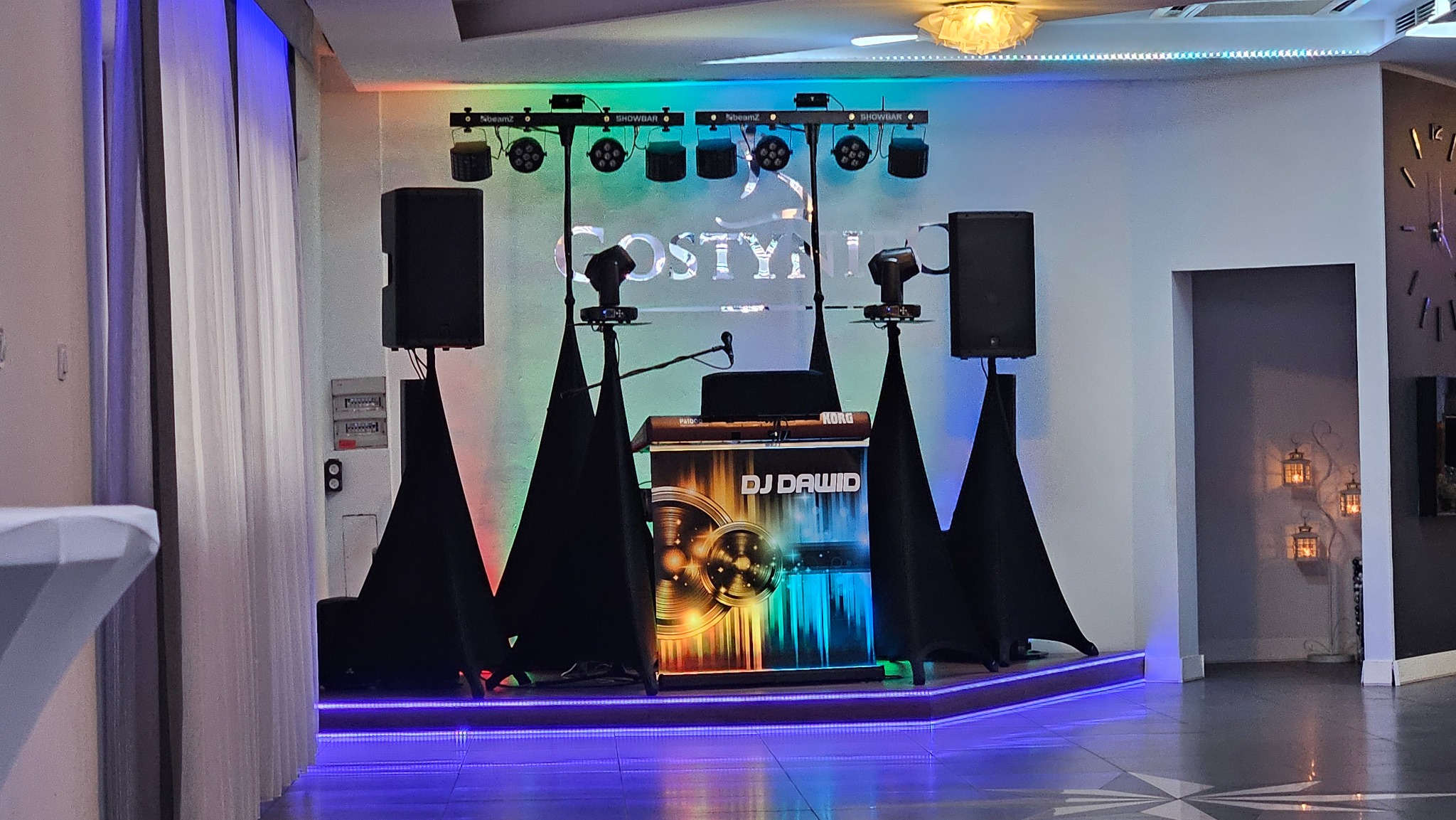 Scena DJ'a Dawida z nagłośnieniem, oświetleniem i konsoletą Korg, gotowa na imprezę w sali z dekoracjami. Podświetlony parkiet i dekoracyjne lampiony w tle.