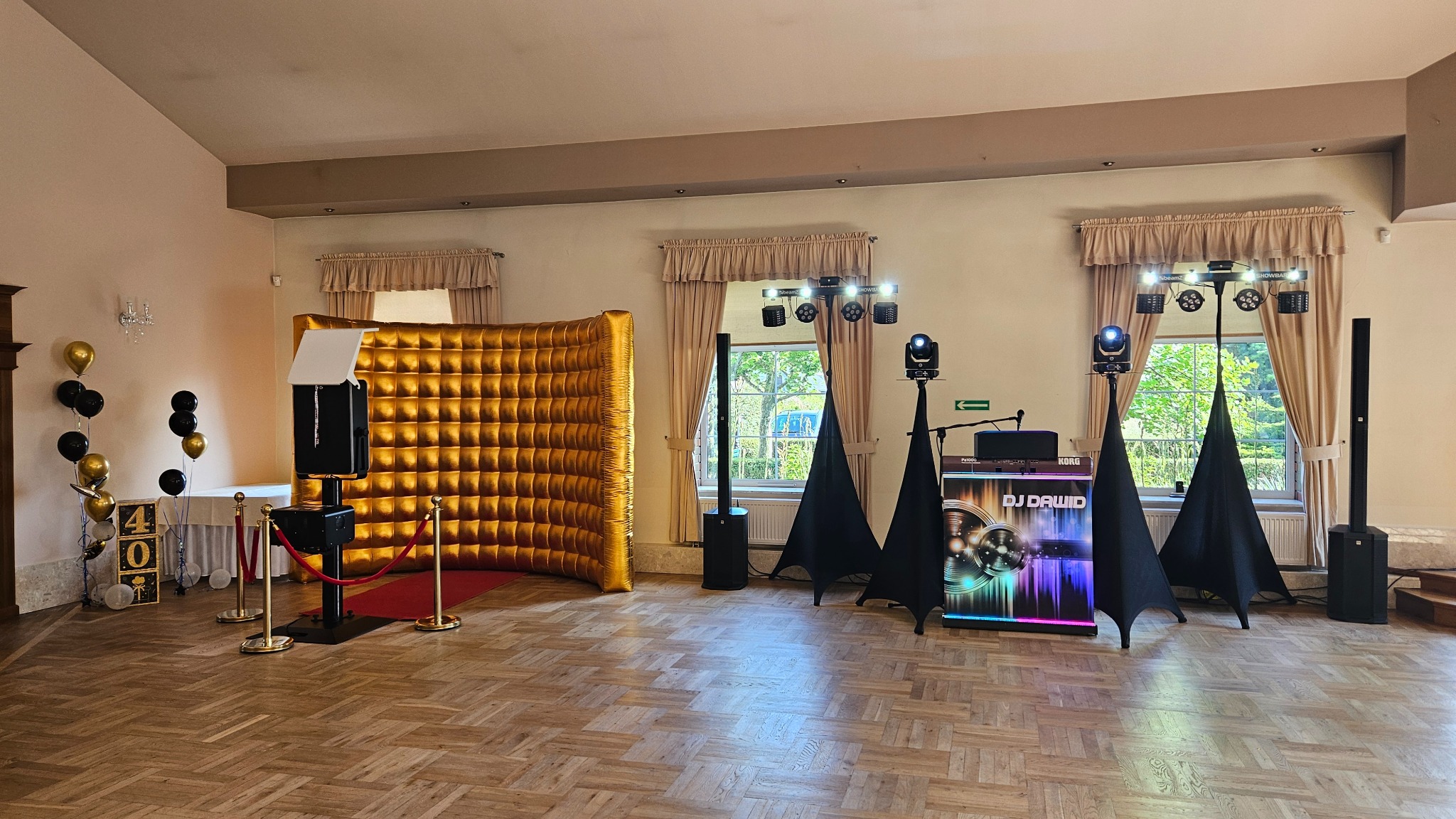 Sala z fotobudką z złotym tłem, nagłośnieniem i oświetleniem. Dekoracje urodzinowe z balonami i napisem '40'. Parkiet w jodełkę, okna z zasłonami.