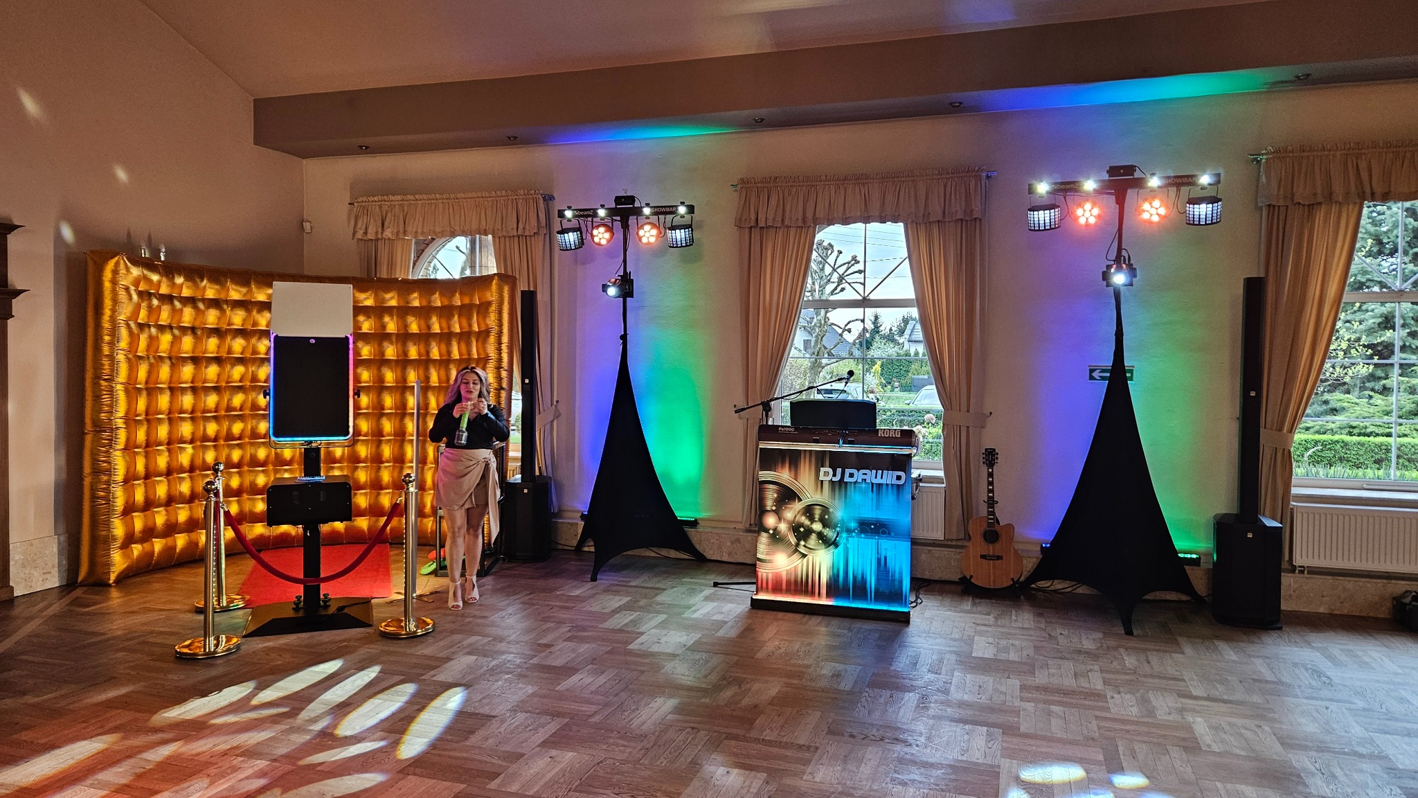 Sala z fotobudką, stanowiskiem DJ'a z napisem 'DJ Dawid', oświetleniem eventowym i osobą trzymającą mikrofon, gotowa na imprezę. Akustyczna gitara oparta o ścianę.
