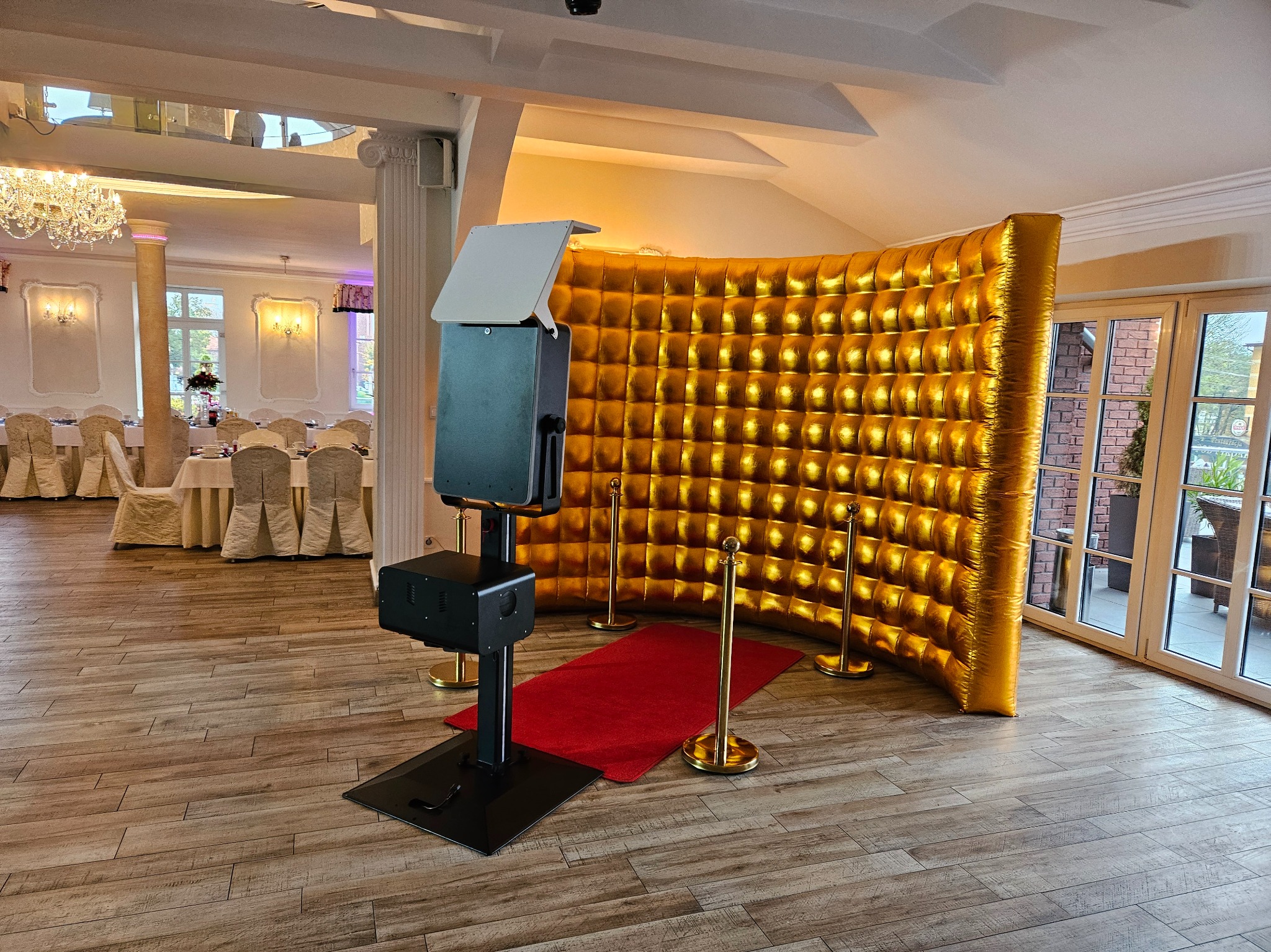 Fotobudka z złotym, dmuchanym tłem w sali bankietowej z parkietem i stołami w tle. Czerwony dywan i złote słupki wyznaczają strefę.