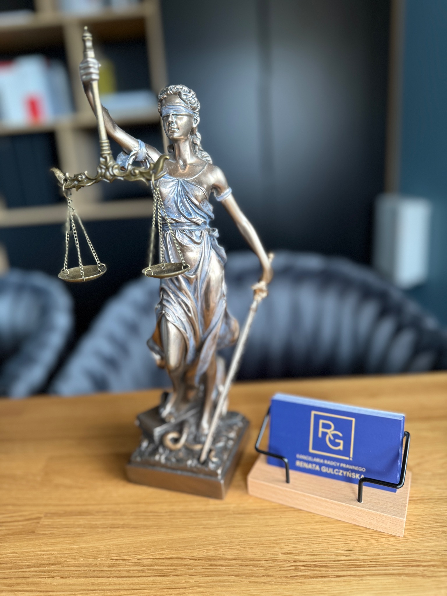 Statuetka Temidy z wagą i mieczem, wizytówka Kancelarii Radcy Prawnego Renaty Gulczyńskiej na drewnianym biurku, w tle regał z książkami i szare krzesła.