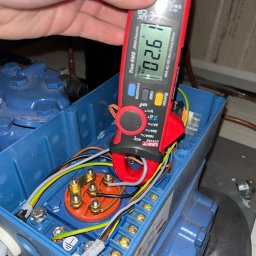 Technika Chłodnicza Jakub Mruk - Pomiar prądu miernikiem cęgowym na niebieskiej skrzynce z przewodami. Widoczny wyświetlacz miernika z wartością 02.61. Detal z bliska, oświetlenie sztuczne.