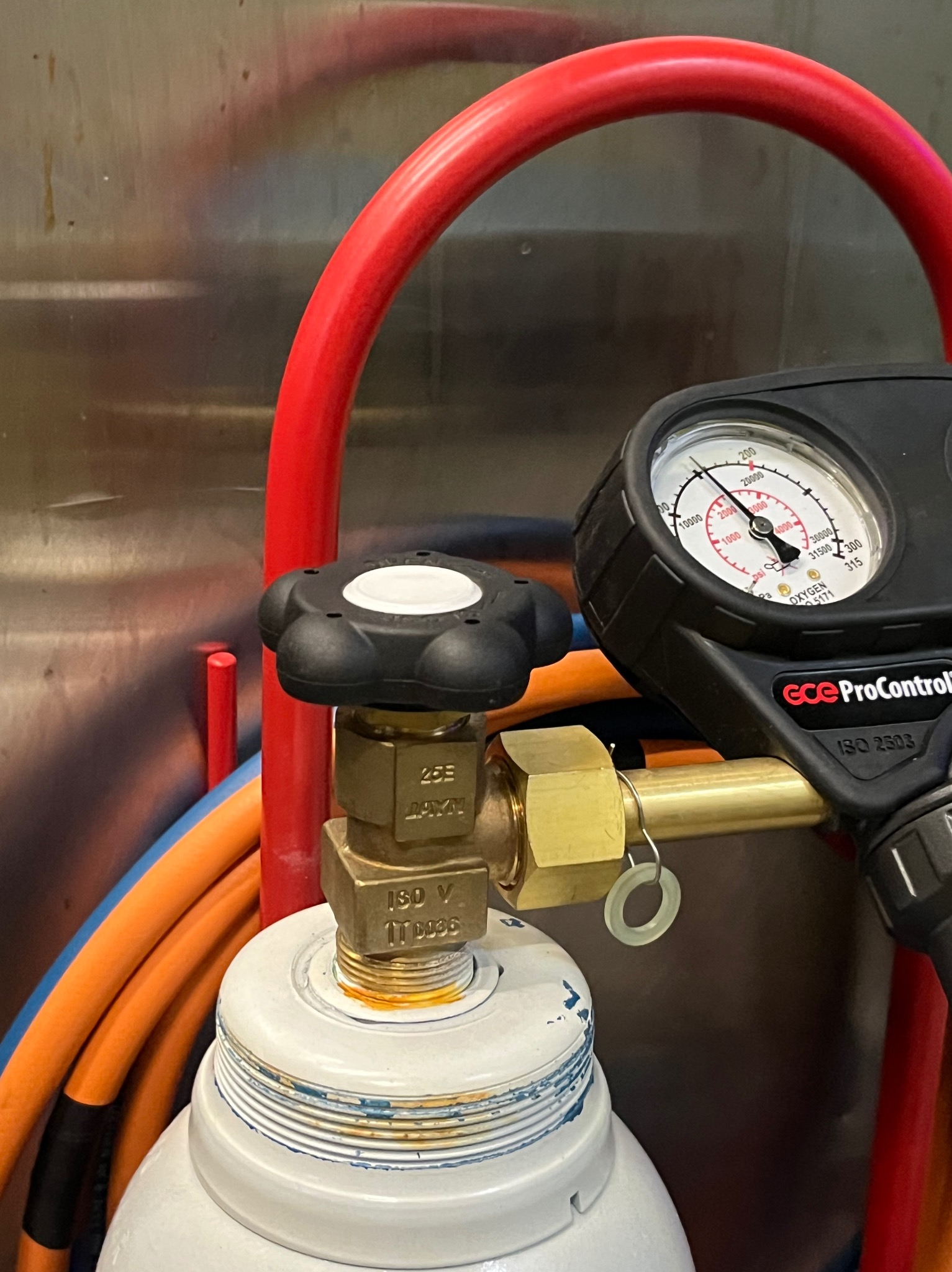 Butla z manometrem i zaworem, widoczne przewody w tle. Sprzęt do pomiaru ciśnienia gazu, prawdopodobnie używany w instalacjach. Detal z bliska, oświetlenie studyjne.