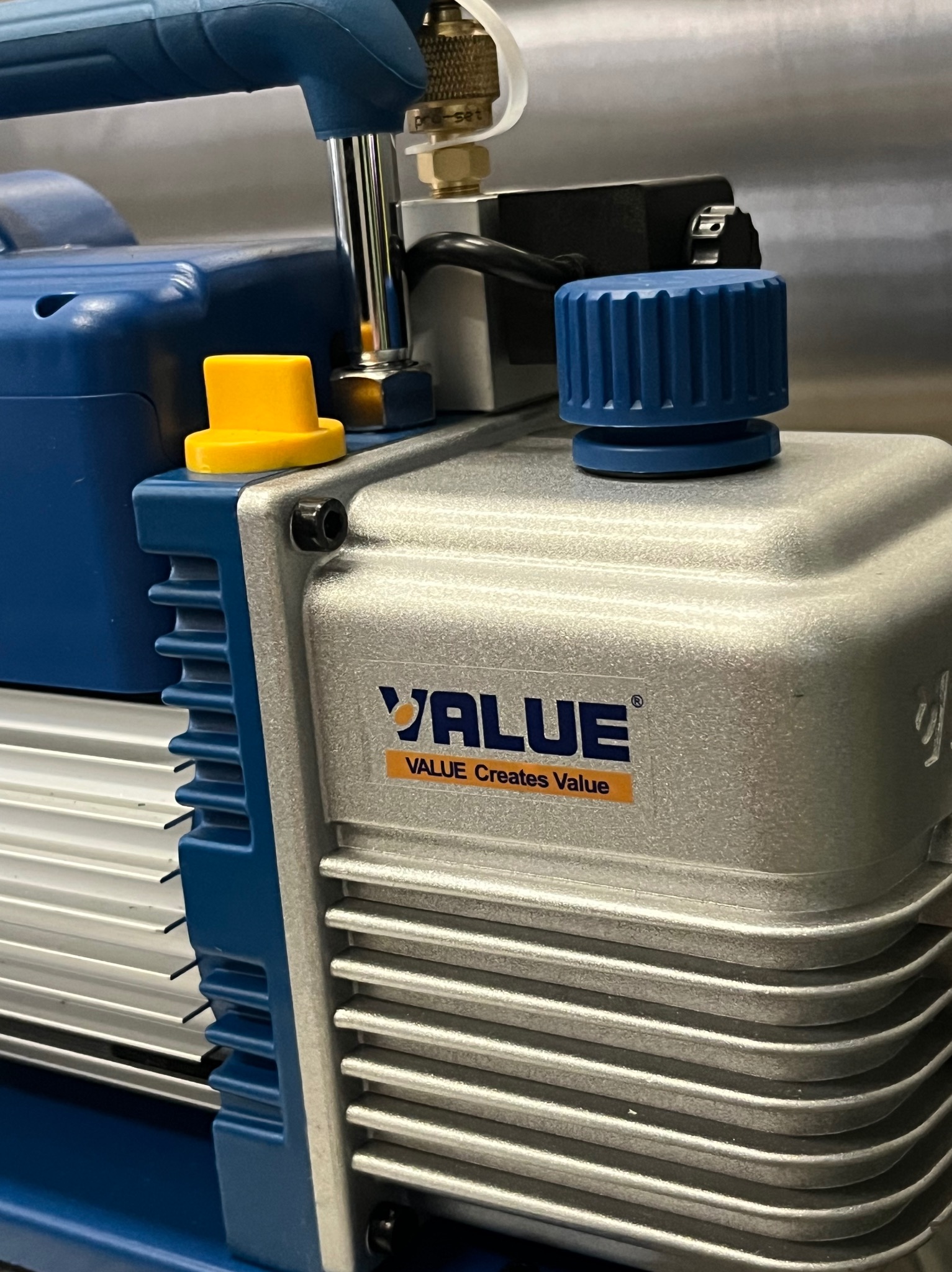 Pompa próżniowa VALUE, srebrno-niebieska z żółtym i niebieskim pokrętłem, widoczne żebra chłodzące i naklejka z logo. Sprzęt do serwisu klimatyzacji.