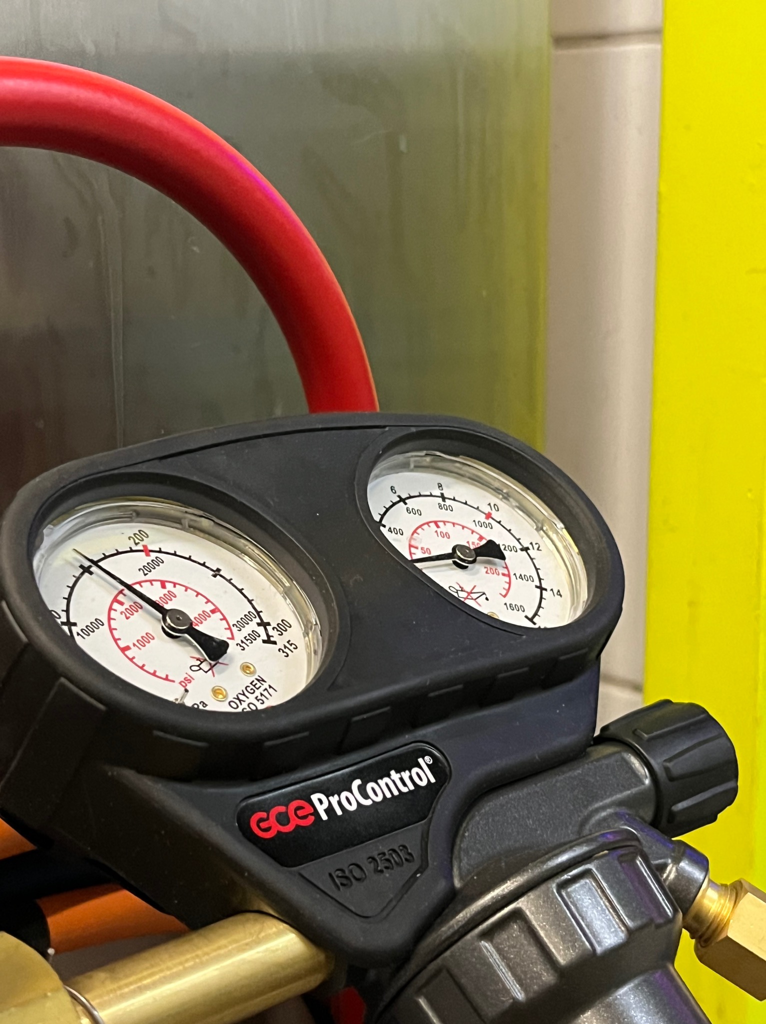 Manometr GCE ProControl z czerwonym wężem i żółtym tłem, wskazujący ciśnienie tlenu. Widoczne wartości PSI i oznaczenia ISO 2503. Detal urządzenia pomiarowego.