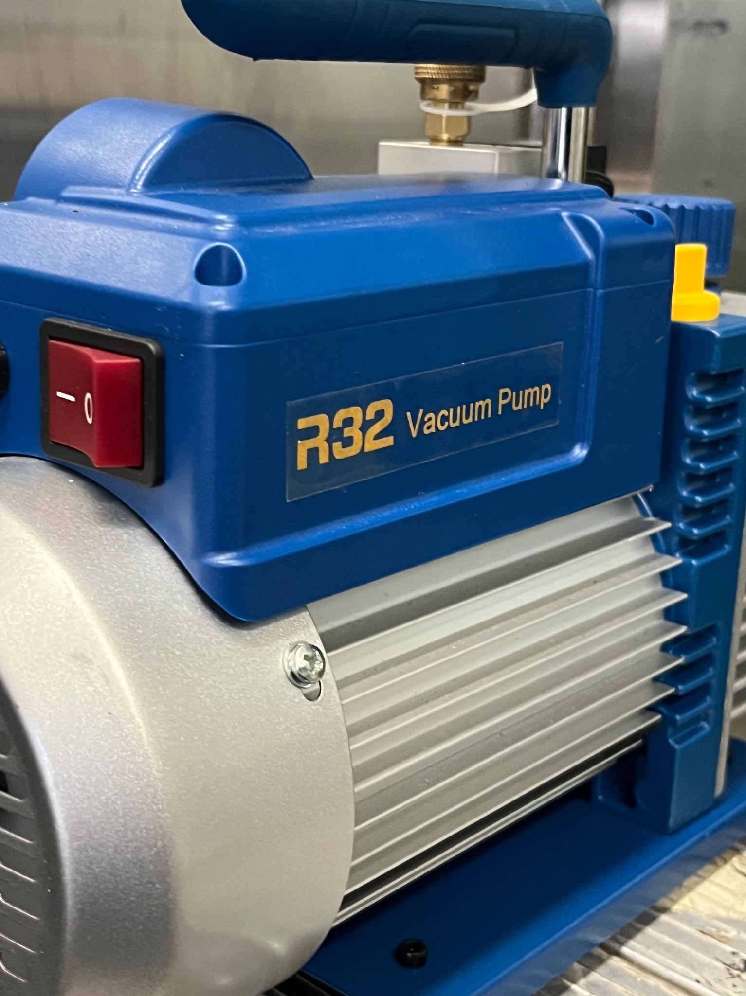 Niebiesko-srebrna pompa próżniowa R32 z czerwonym włącznikiem, widoczna z bliska, z metalowymi żebrami chłodzącymi i elementami złącznymi.