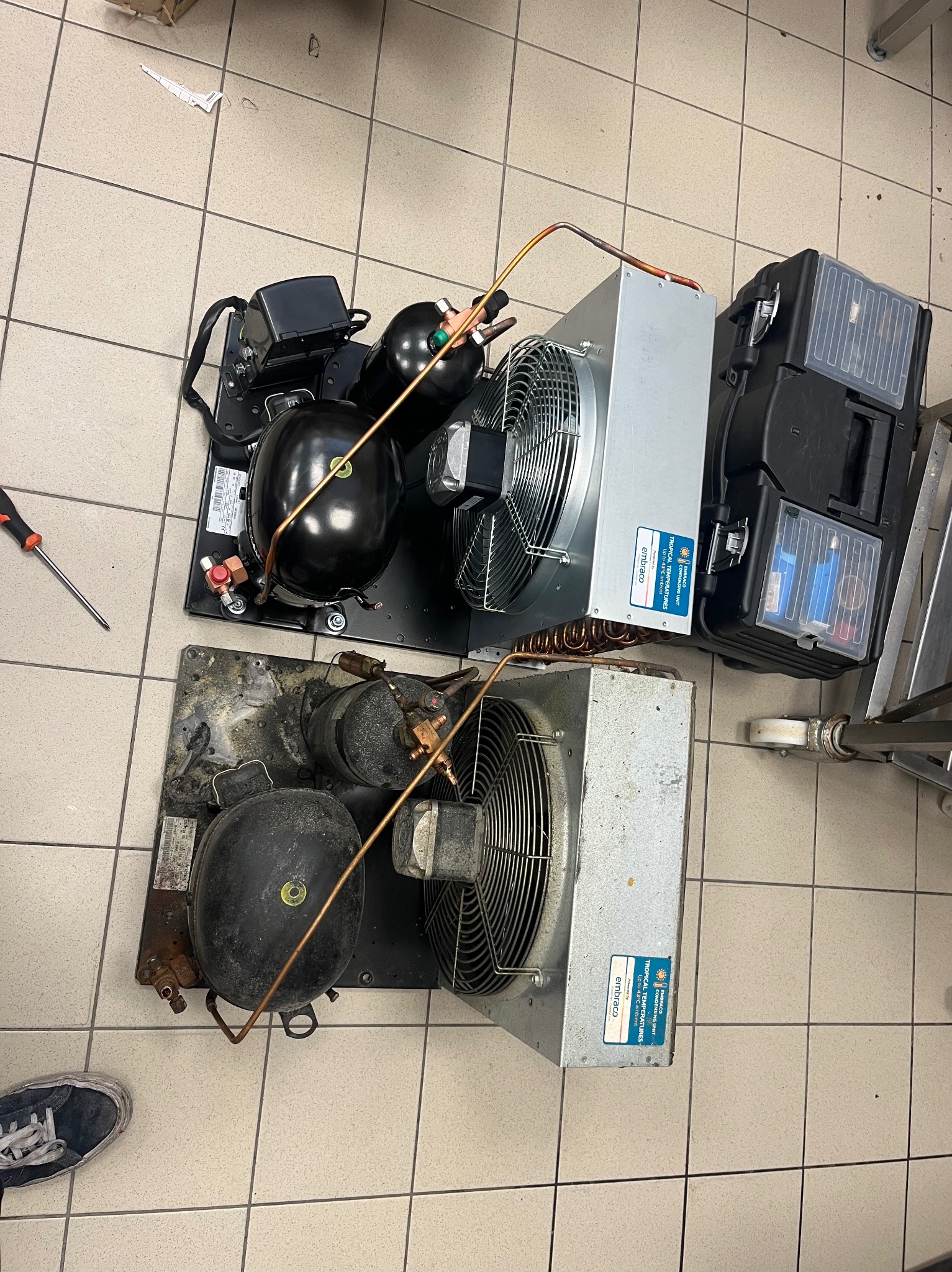 Dwa agregaty chłodnicze na podłodze z płytek, jeden nowy, drugi używany, obok skrzynka narzędziowa i śrubokręt. Widoczne detale konstrukcyjne i oznaczenia producenta.