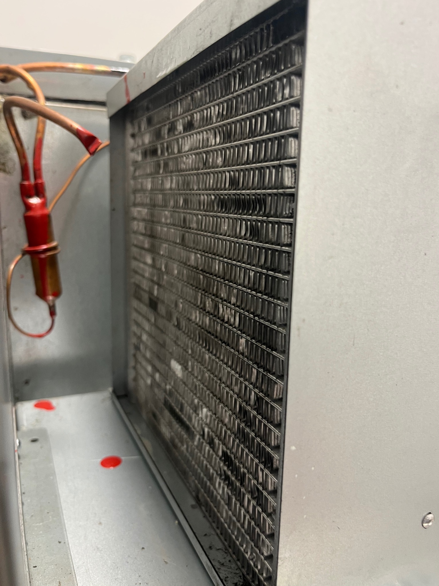 Zabrudzony wymiennik ciepła w metalowej obudowie, widoczne rurki miedziane z elementami czerwonymi. Detal zanieczyszczonej powierzchni i czerwone plamy na spodzie.