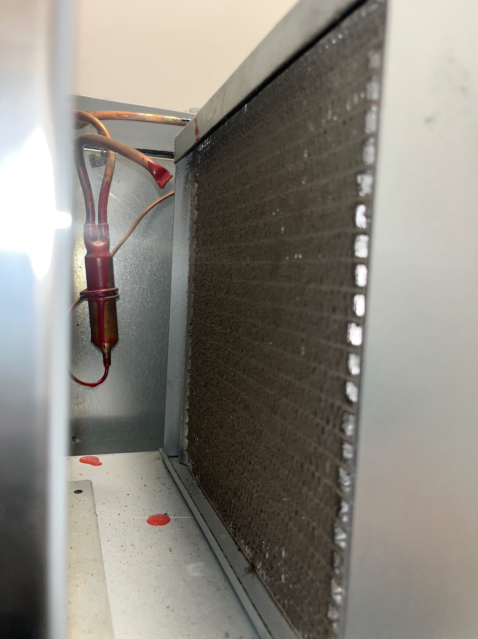 Zanieczyszczony filtr klimatyzacji z widocznymi elementami instalacji chłodniczej, wewnątrz obudowy. Widoczne ślady czerwonej substancji na elementach i podłożu.