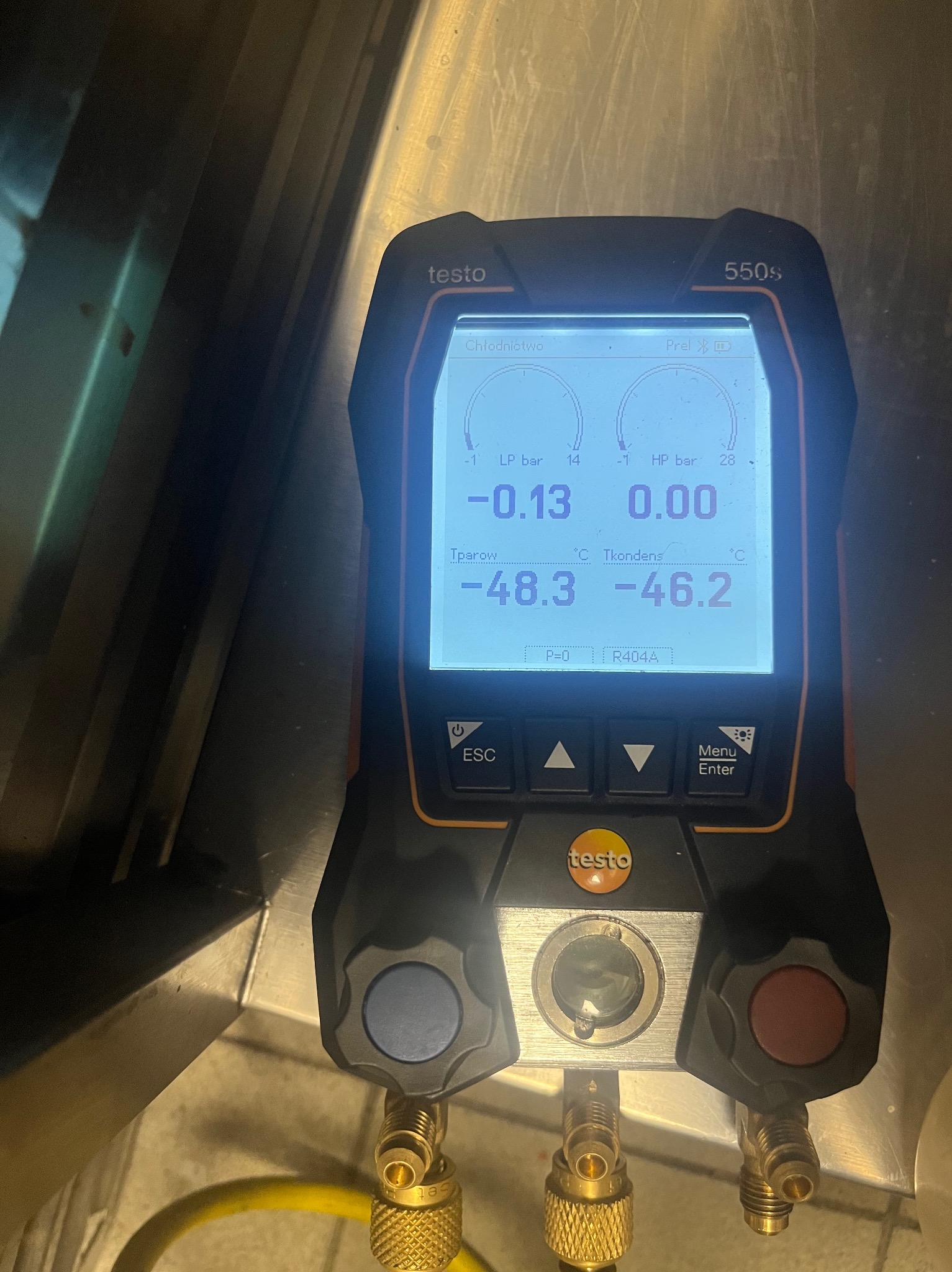 Miernik cyfrowy Testo 550s z podłączonymi przewodami, wyświetlający parametry chłodnicze, takie jak ciśnienie i temperatura, na tle metalowej powierzchni.