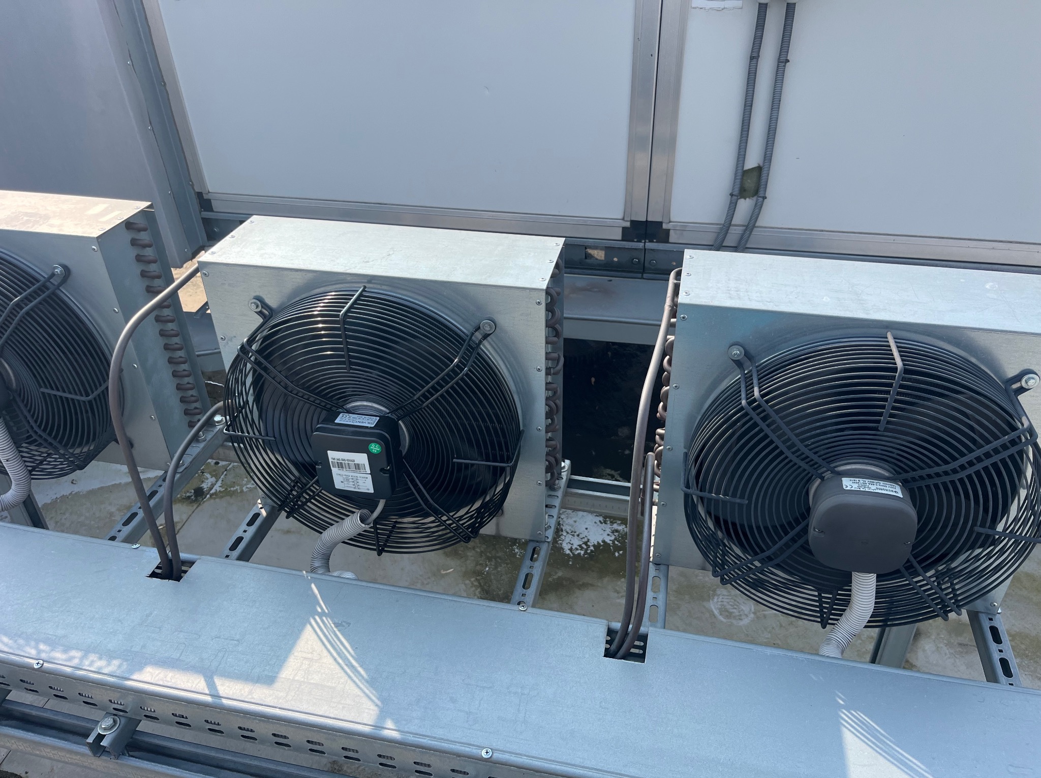 Zewnętrzne jednostki wentylacyjne na dachu, z widocznymi wentylatorami i metalową konstrukcją wsporczą. Szare przewody prowadzą do urządzeń. Widok z góry, oświetlenie dzienne.