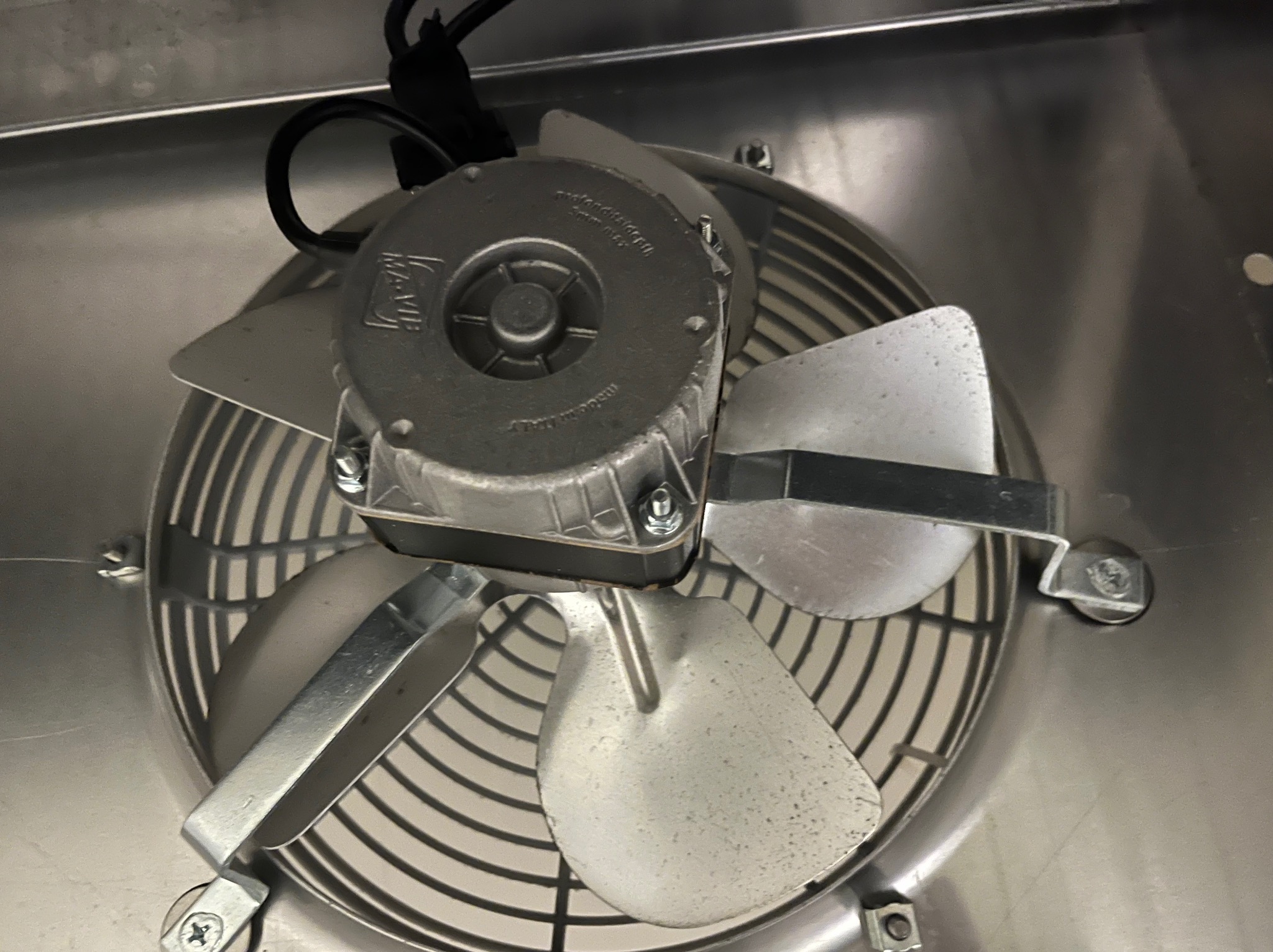 Metalowy wentylator z trzema łopatkami w obudowie, widoczny z góry, zamontowany w metalowej konstrukcji. Widoczne śruby mocujące i przewody.