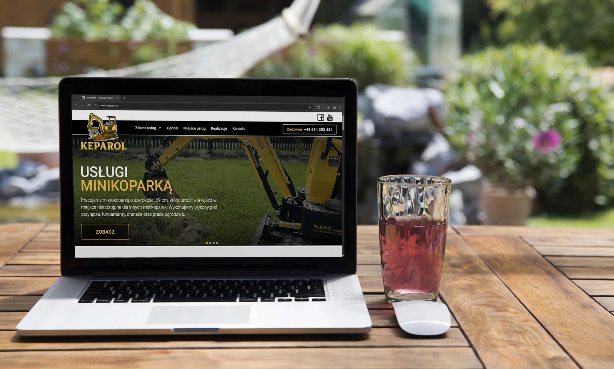 Laptop na drewnianym stole prezentuje stronę internetową firmy Keparol z ofertą usług minikoparką. W tle zieleń ogrodu, obok szklanka z napojem i myszka.