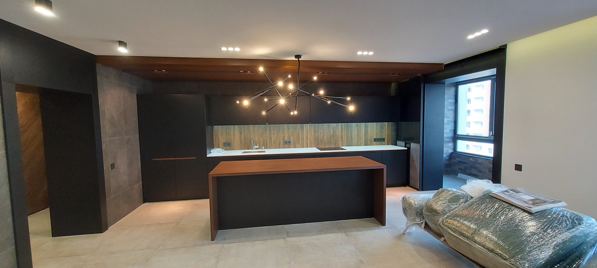 Nowoczesna kuchnia z wyspą, czarne szafki, drewniane akcenty na suficie i ścianie, designerska lampa. Częściowo zakryte meble folią ochronną.