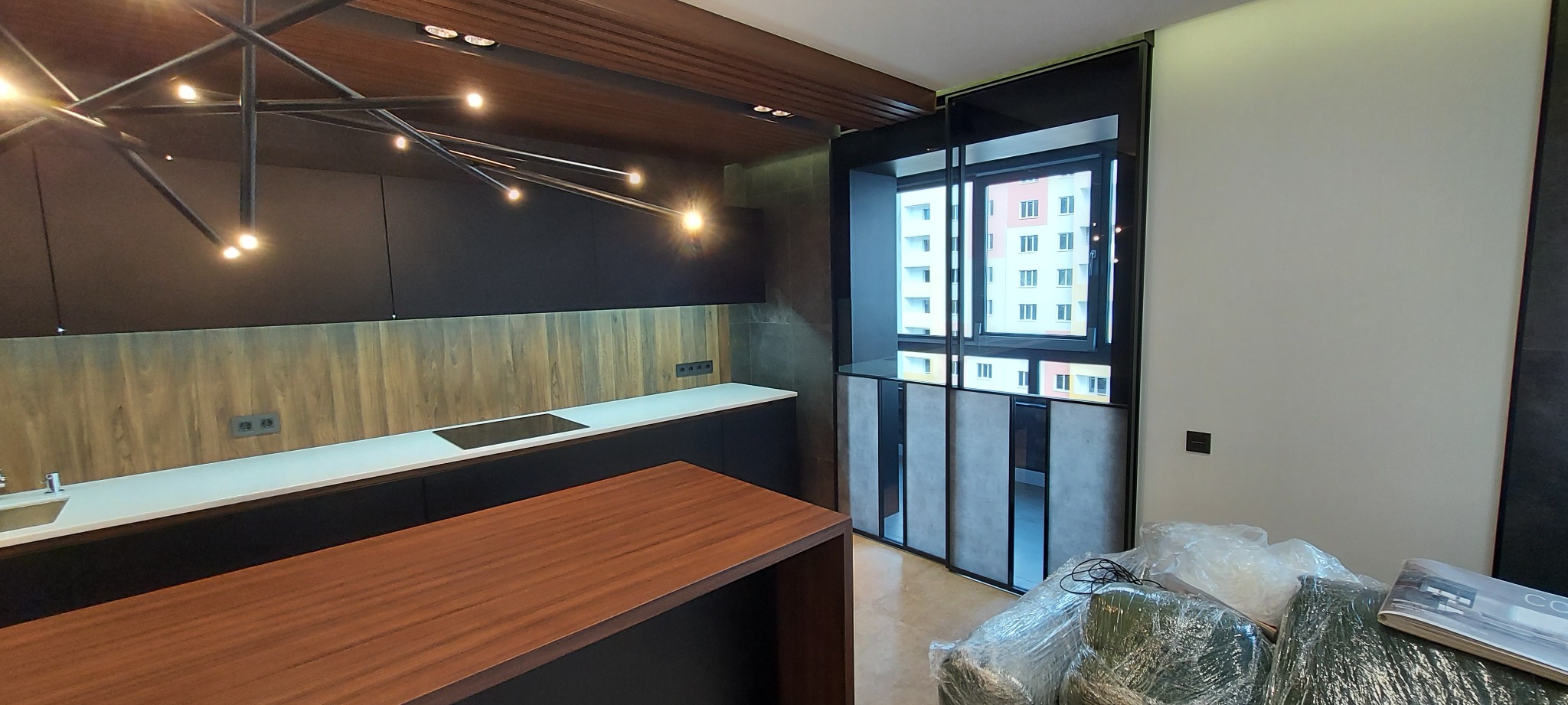 Nowoczesna kuchnia z ciemnymi szafkami, drewnianym panelem i wyspą. Widoczne okno balkonowe i designerska lampa. Częściowo zakryte meble folią ochronną.