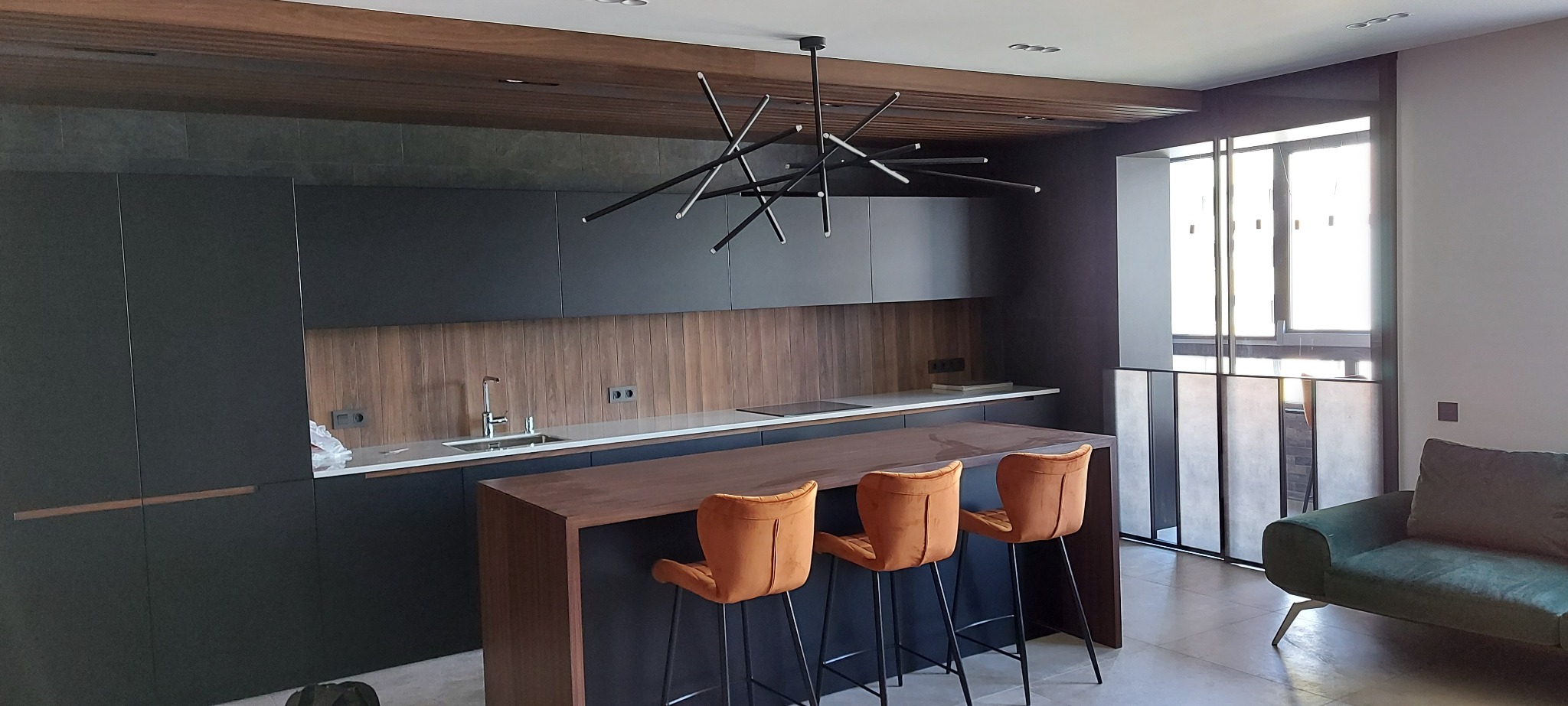 Nowoczesna kuchnia z wyspą i hokerami w odcieniach brązu i czerni. Drewniane wykończenie ściany nad blatem. Designerska lampa sufitowa. Minimalistyczny styl.