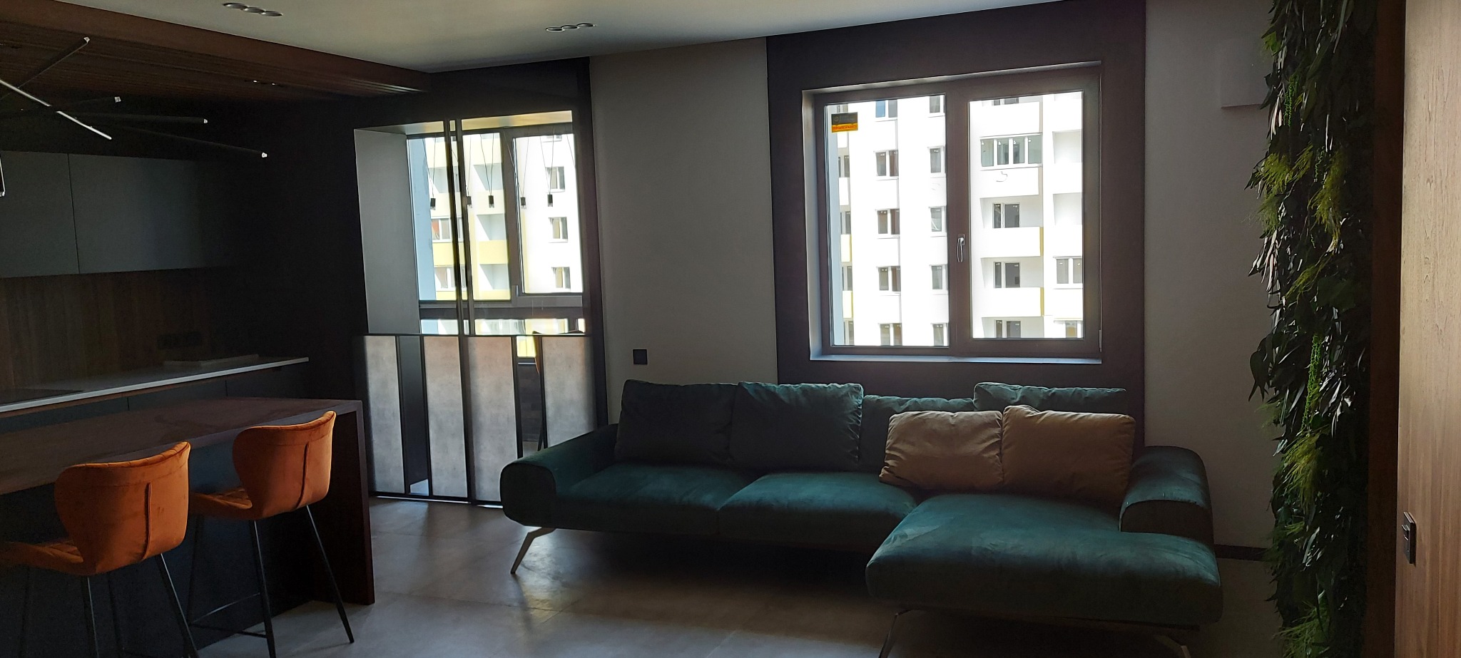 Nowoczesne wnętrze: zielona sofa, pomarańczowe krzesła barowe, ściana z roślin i okno z widokiem na budynek. Minimalistyczny design z akcentami kolorystycznymi.