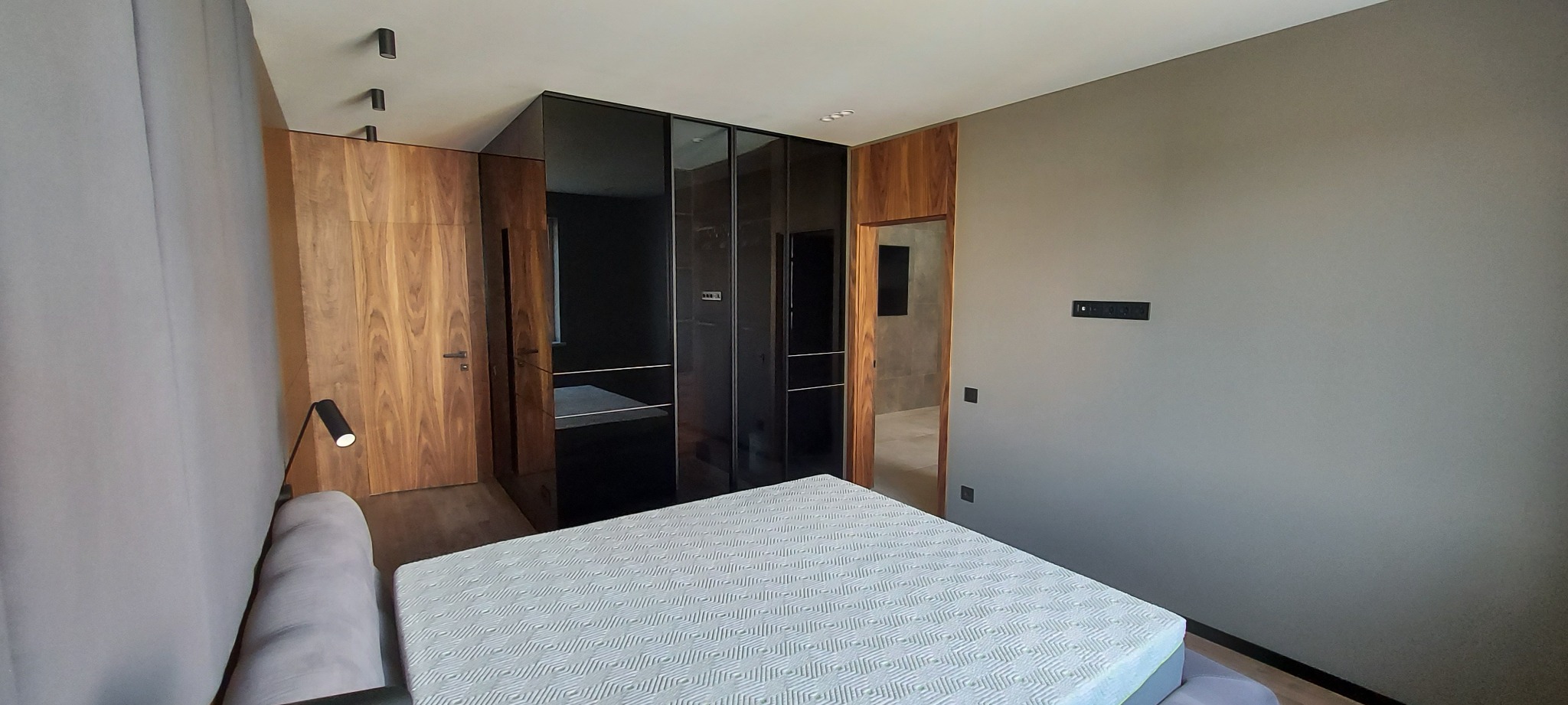 Nowoczesna sypialnia z łóżkiem, zabudową z ciemnego szkła i drewnianymi elementami. Minimalistyczny design, stonowana kolorystyka, oświetlenie punktowe.