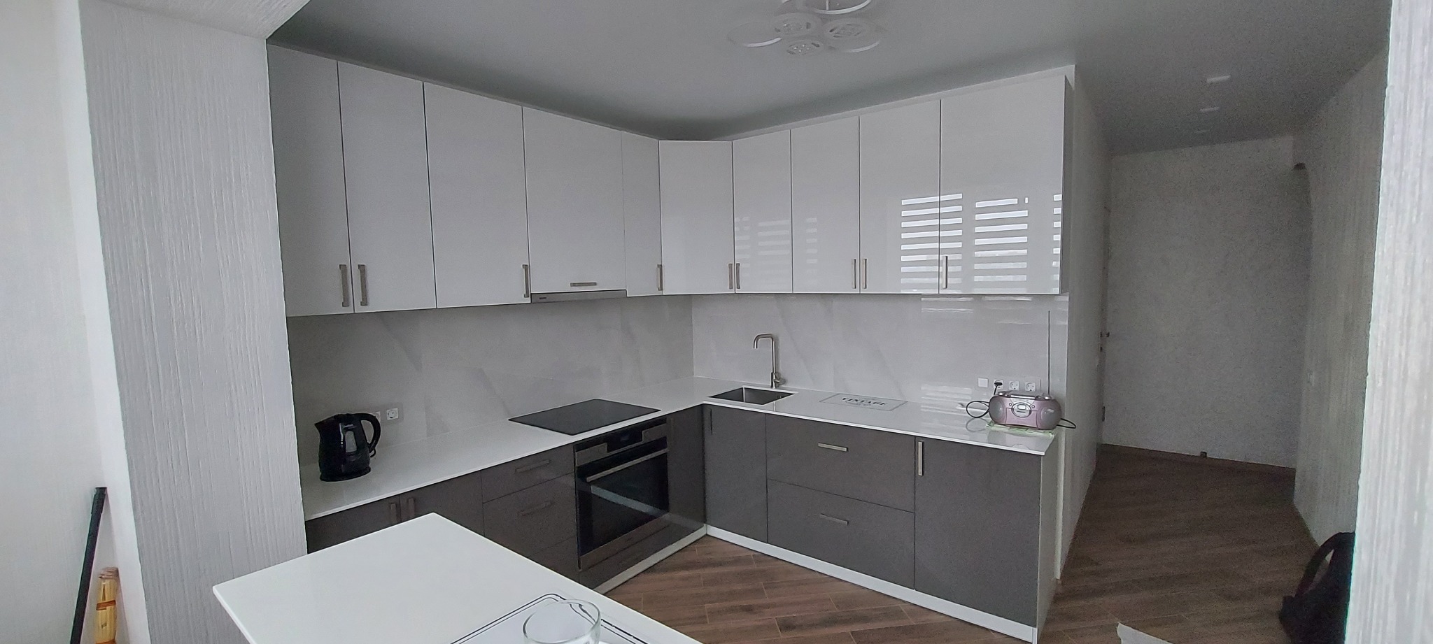 Nowoczesna kuchnia w odcieniach bieli i szarości z błyszczącymi frontami, wyposażona w płytę indukcyjną i piekarnik. Widoczny czajnik elektryczny i radio w stylu retro.