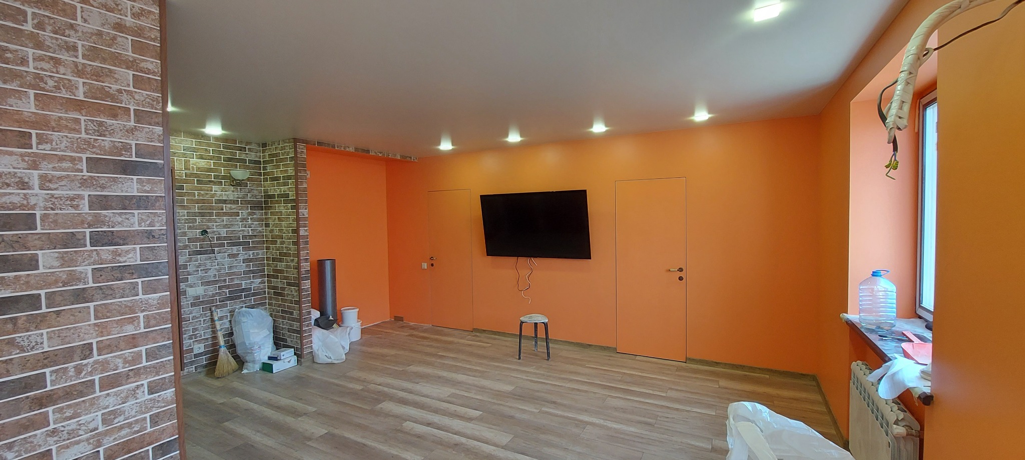 Pomarańczowe ściany, panele podłogowe i dekoracyjna ściana z cegły w nowocześnie wykończonym pokoju z telewizorem i ukrytymi drzwiami.