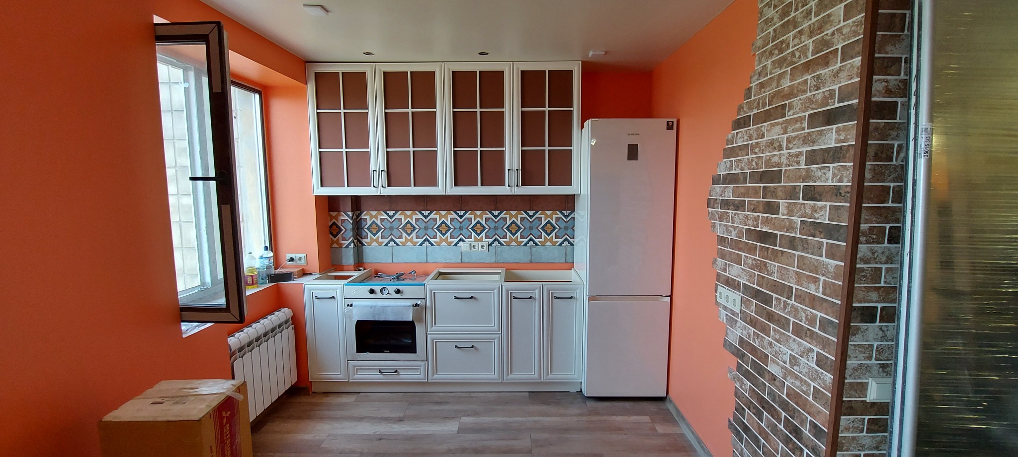Jasna kuchnia z białymi szafkami i ceglaną ścianą w Częstochowie. Widoczne okno, grzejnik i sprzęt AGD. Ściany pomalowane na pomarańczowo.