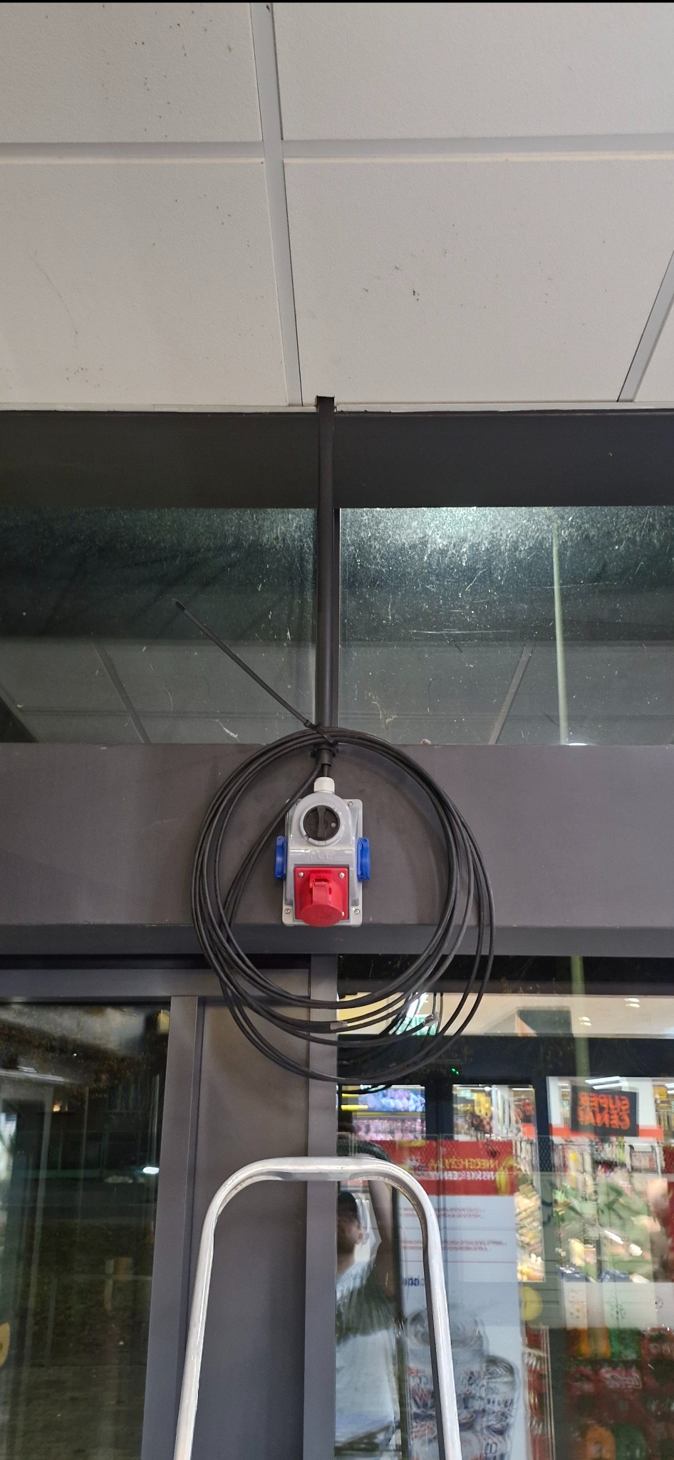 Zamontowane gniazdo siłowe z przewodem zwisającym w pętli, widoczne na tle sufitu i okna wystawowego sklepu. Instalacja elektryczna w obiekcie handlowym.