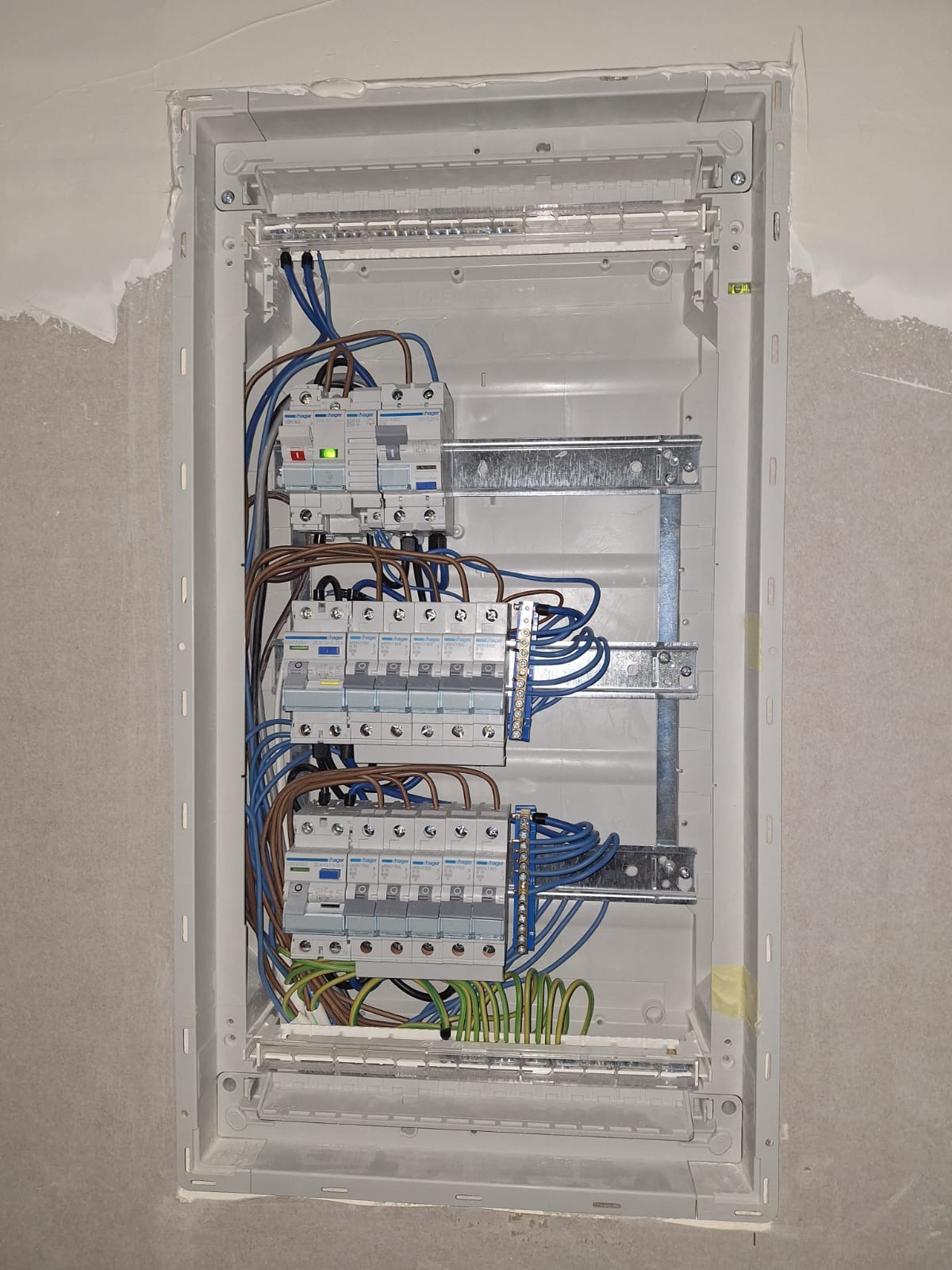 Wnętrze skrzynki rozdzielczej z zamontowanymi bezpiecznikami Hager, okablowaniem w kolorach niebieskim, brązowym i zielono-żółtym. Widoczne szyny montażowe i poziomica.