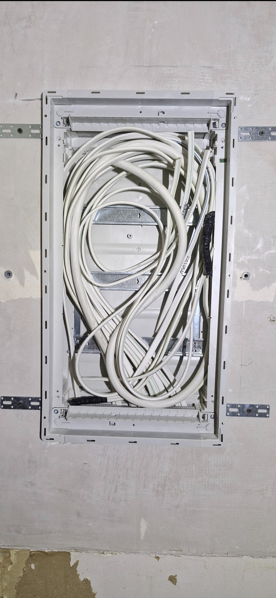 Wewnętrzny widok skrzynki elektrycznej w ścianie z licznymi białymi kablami, częściowo owiniętymi czarną taśmą. Widoczne metalowe elementy montażowe.