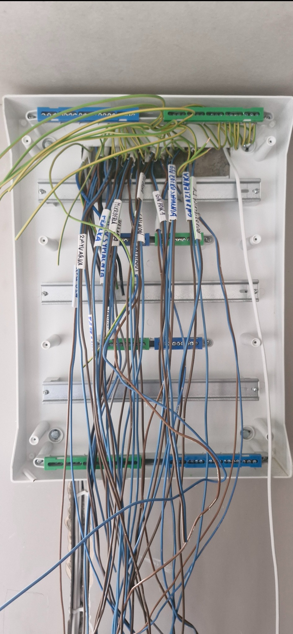 Rozdzielnia elektryczna w białej obudowie z oznaczonymi przewodami (Zmywarka, Sypialnia, Mikrofala, Salon, Łazienka). Widoczne listwy zaciskowe i szyny montażowe.