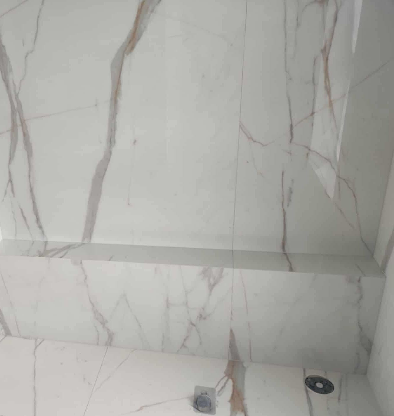Białe płytki imitujące marmur z szarymi i złotymi żyłami, ułożone na ścianie i podłodze łazienki, widoczne z perspektywy pod kątem, z odpływem liniowym w rogu.