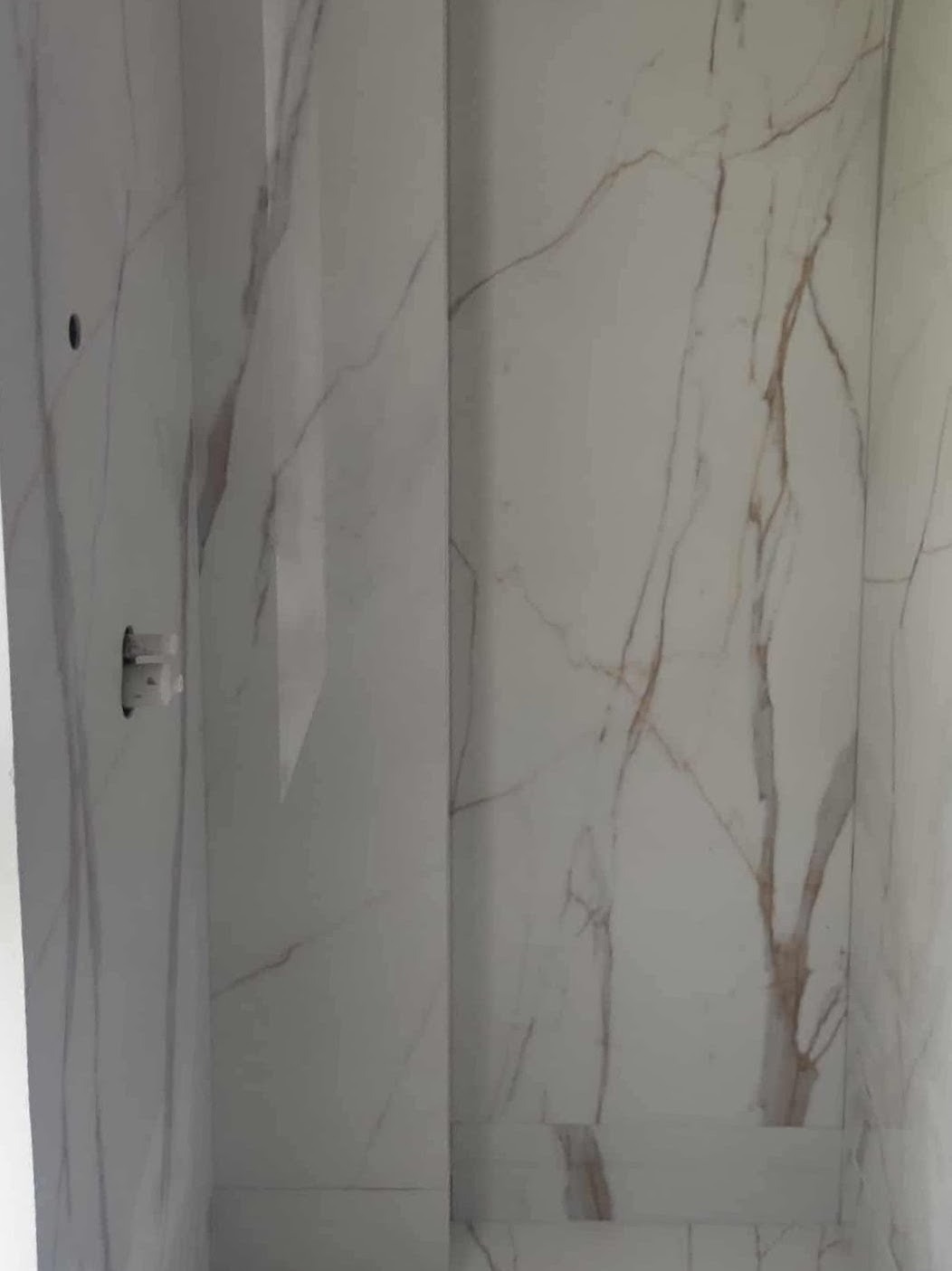 Wykończona ściana w łazience z białych płyt imitujących marmur z brązowymi żyłami, widoczny otwór na baterię. Realizacja glazurnicza w Słupsku.