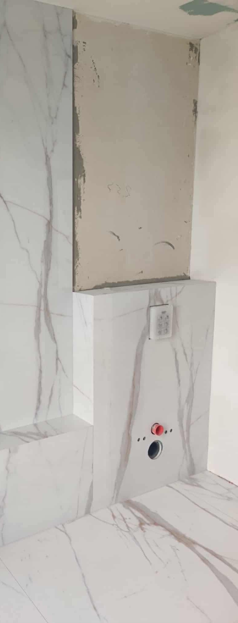 Biała marmurowa okładzina ściany z widocznymi otworami na instalacje i fragmentem surowej ściany, ukazująca etap prac wykończeniowych w łazience.