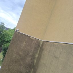 Kolor wash - Częściowo odnowiona elewacja budynku z widoczną różnicą w kolorze i czystości, oddzielona listwą. U góry jasny beż, dół ciemniejszy, zabrudzony. Widoczna kamera.