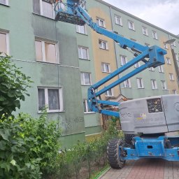 Kolor wash - Pracownik na podnośniku czyści elewację budynku myjką ciśnieniową. Widoczny efekt czyszczenia - różnica w kolorze elewacji. Budynek wielorodzinny, zieleń w tle.