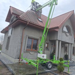 Kolor wash - Pracownik w żółtym kombinezonie na podnośniku koszowym czyści dach domu z czerwonej dachówki. Widoczna szara elewacja budynku i zielony podnośnik.