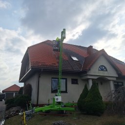 Kolor wash - Pracownik na podnośniku koszowym czyści dach domu jednorodzinnego. Widoczna część dachu przed i po czyszczeniu, z wyraźną różnicą w kolorze. Pochmurne niebo w tle.