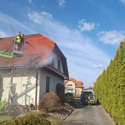 Kolor wash - Pracownik w żółtej kamizelce, na podnośniku, czyści dach domu z czerwonej dachówki strumieniem wody pod ciśnieniem. Widoczne panele słoneczne i niebieskie niebo.