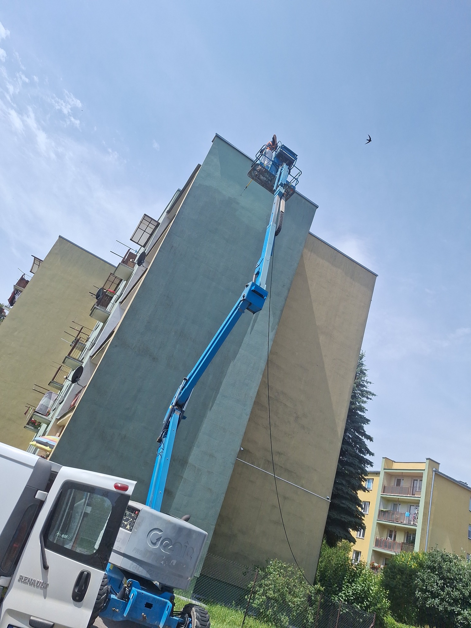 Praca na wysokości: niebieski podnośnik koszowy Genie przy malowaniu bloku. Widoczny fragment białego samochodu dostawczego i sylwetka ptaka na tle błękitnego nieba.