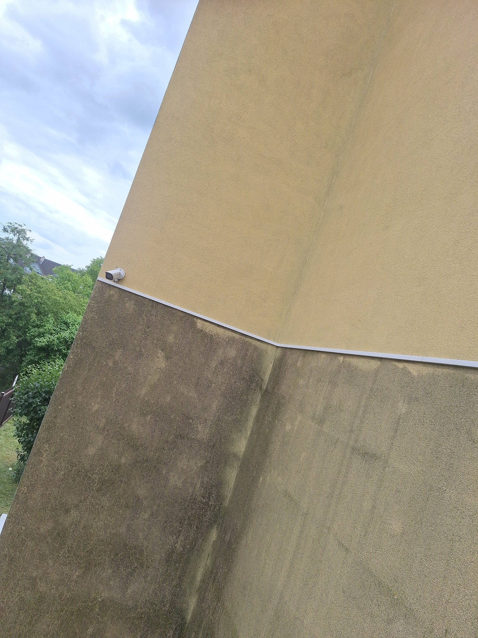 Częściowo odnowiona elewacja budynku z widoczną różnicą w kolorze i czystości, oddzielona listwą. U góry jasny beż, dół ciemniejszy, zabrudzony. Widoczna kamera.