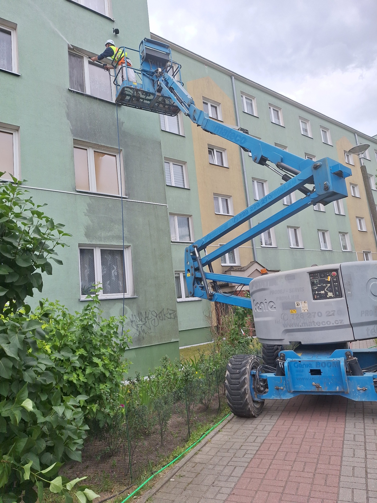 Pracownik na podnośniku czyści elewację budynku myjką ciśnieniową. Widoczny efekt czyszczenia - różnica w kolorze elewacji. Budynek wielorodzinny, zieleń w tle.