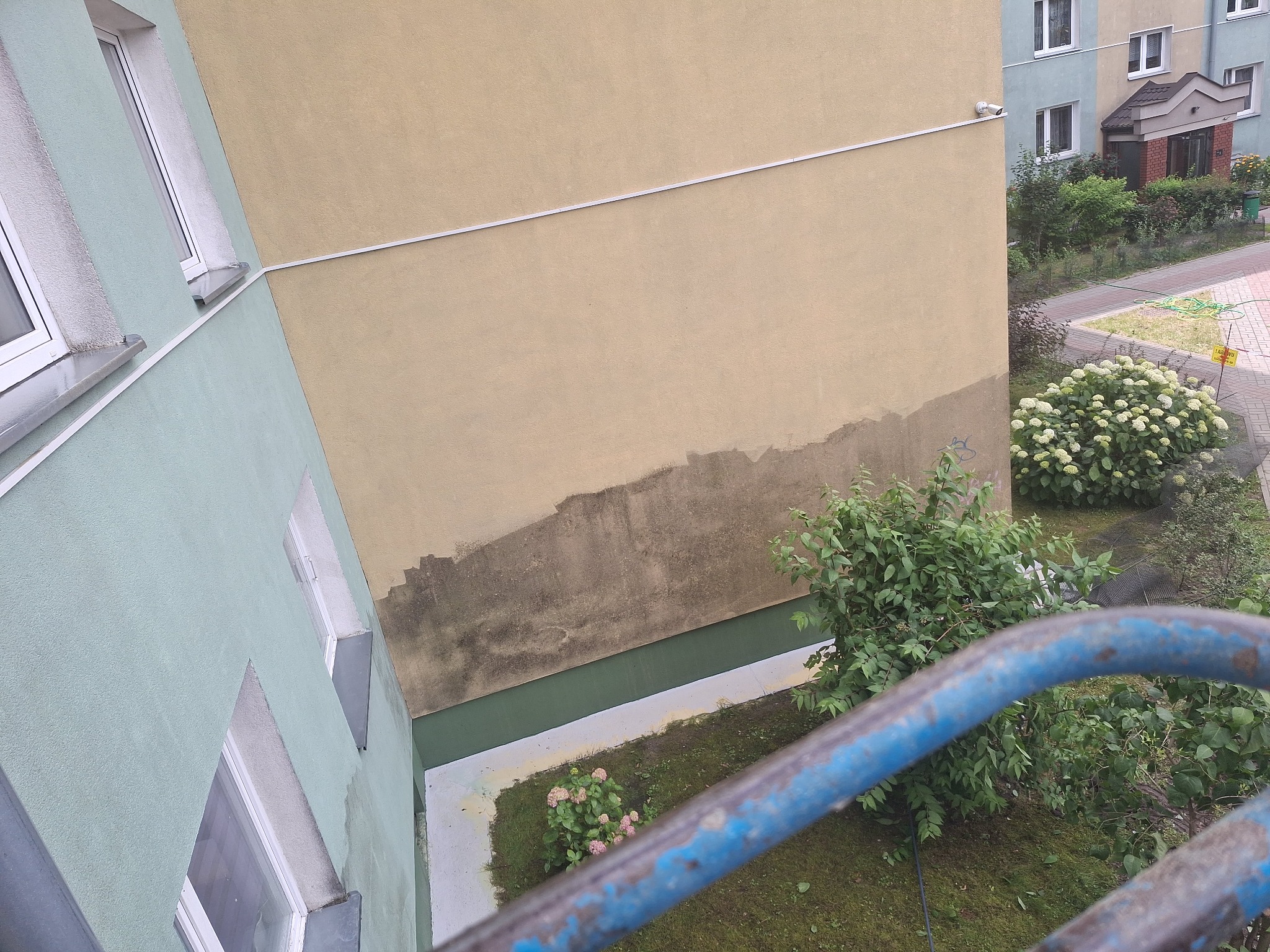 Fragment elewacji budynku z widocznymi śladami wilgoci i zabrudzeń przed planowanym malowaniem. Widok z góry, z fragmentem niebieskiej barierki w kadrze.