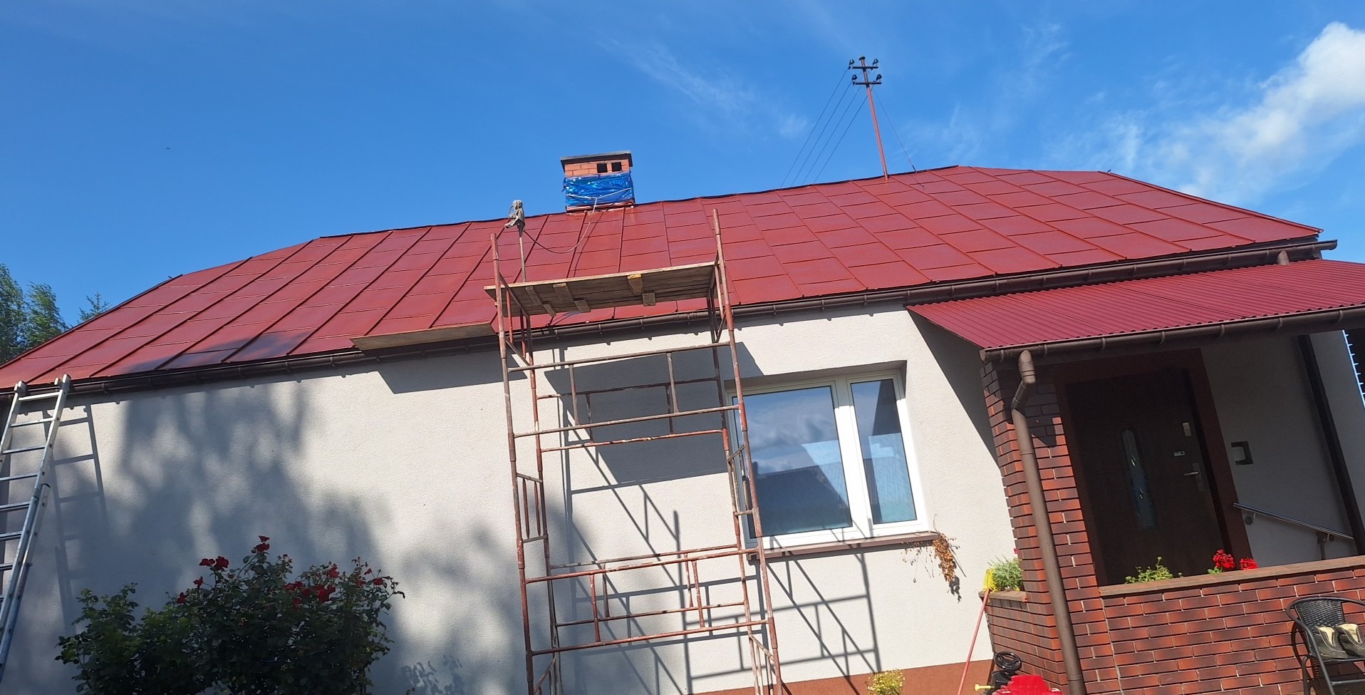 Dom jednorodzinny z nowym, czerwonym dachem. Widoczne rusztowanie i drabina, sugerujące prace dekarskie. Błękitne niebo w tle.
