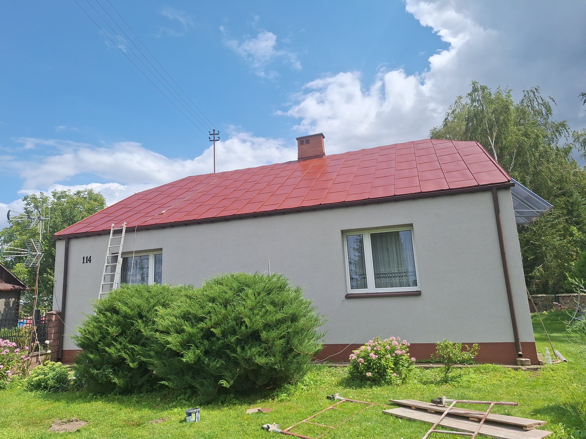 Dom z czerwoną dachówką w Białej Podlaskiej po renowacji. Świeżo pomalowana elewacja, drabina oparta o ścianę, zadbany ogród. Słoneczny dzień.