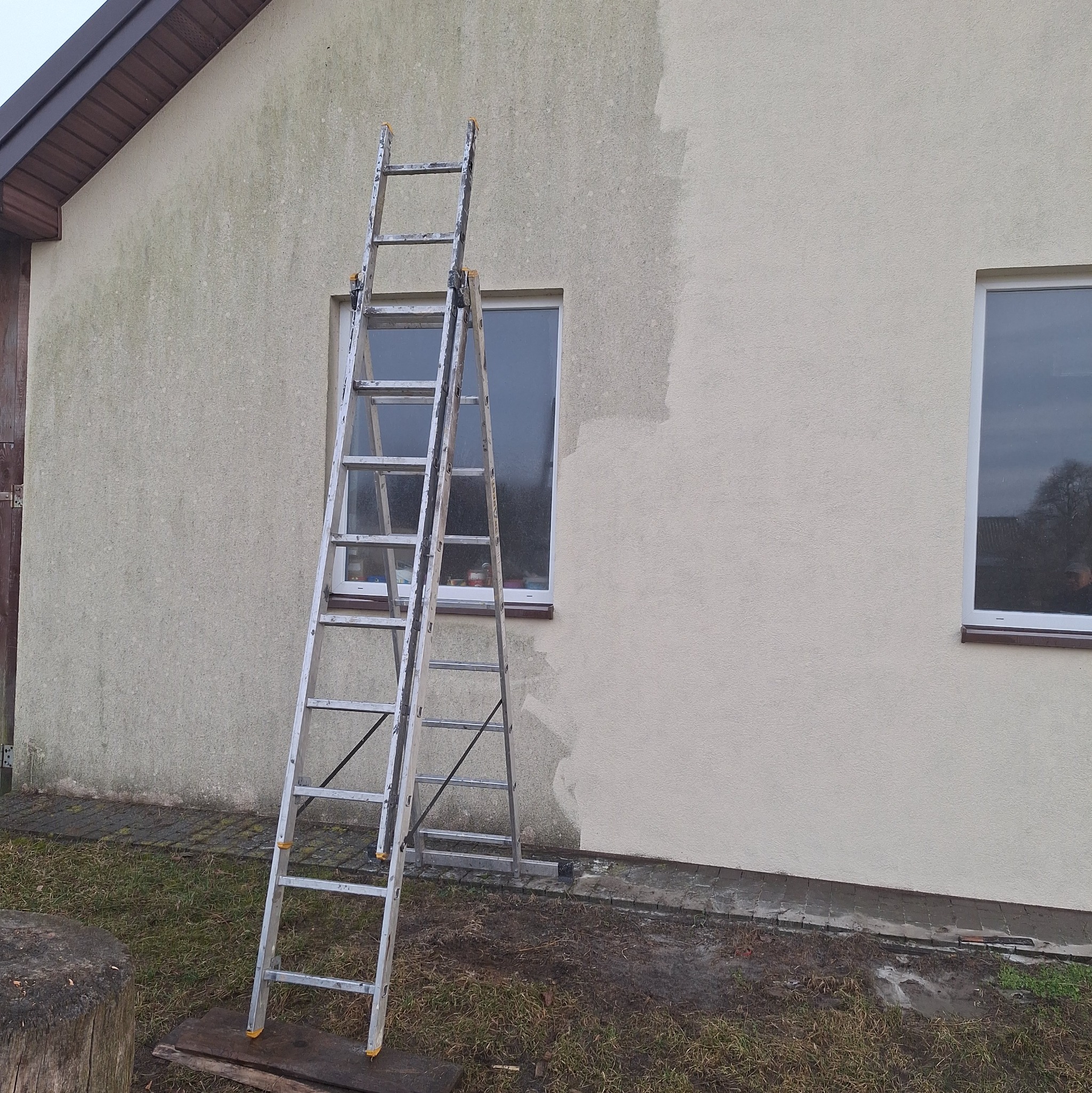 Fragment elewacji domu z drabiną, ukazujący obszar po malowaniu i obszar wymagający odświeżenia. Widoczne różnice w kolorze i czystości powierzchni.