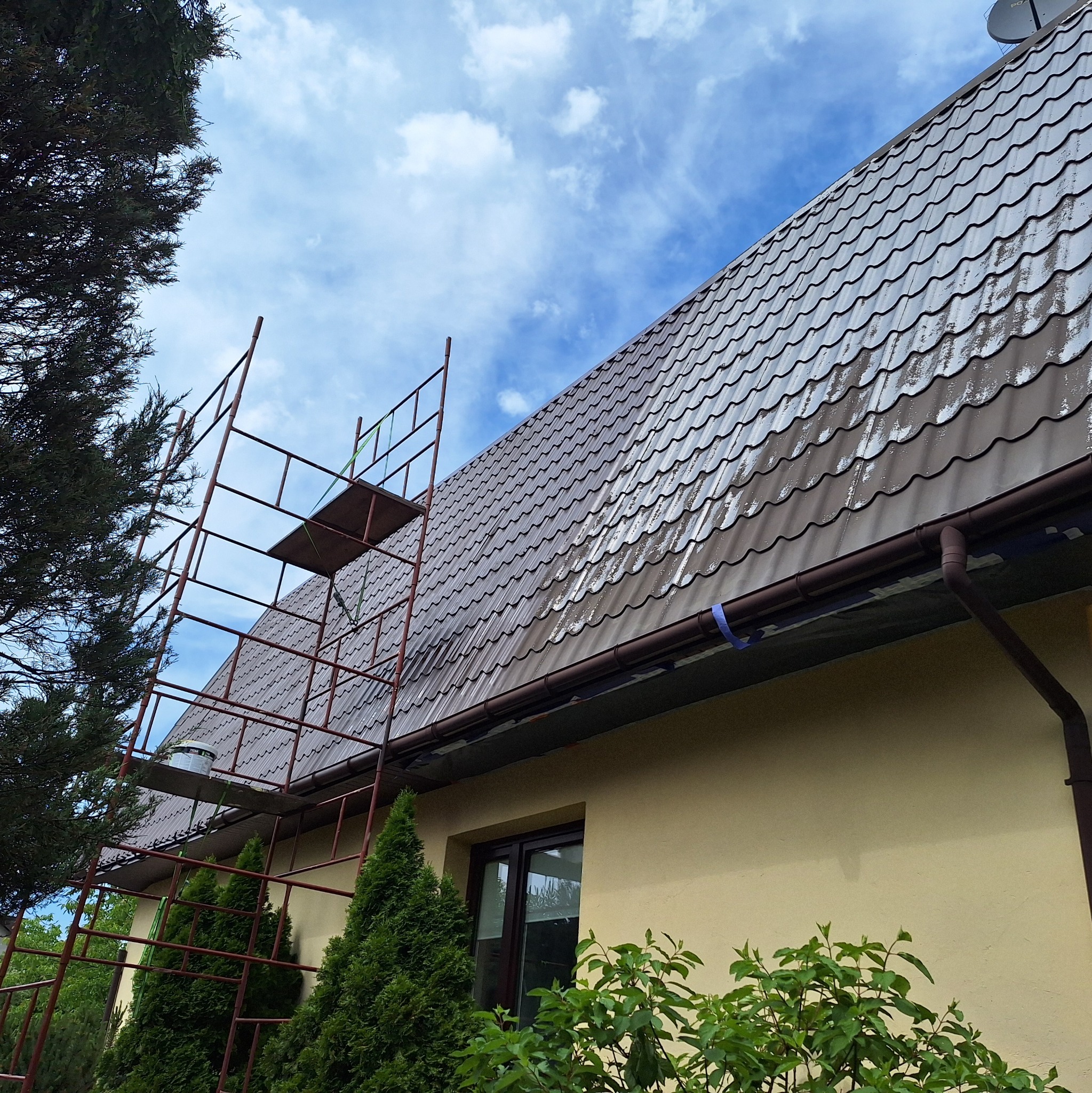 Malowanie dachu domu jednorodzinnego z użyciem rusztowania. Widoczny fragment elewacji i roślinność. Dachówka metalowa, częściowo pomalowana, z widocznymi smugami.