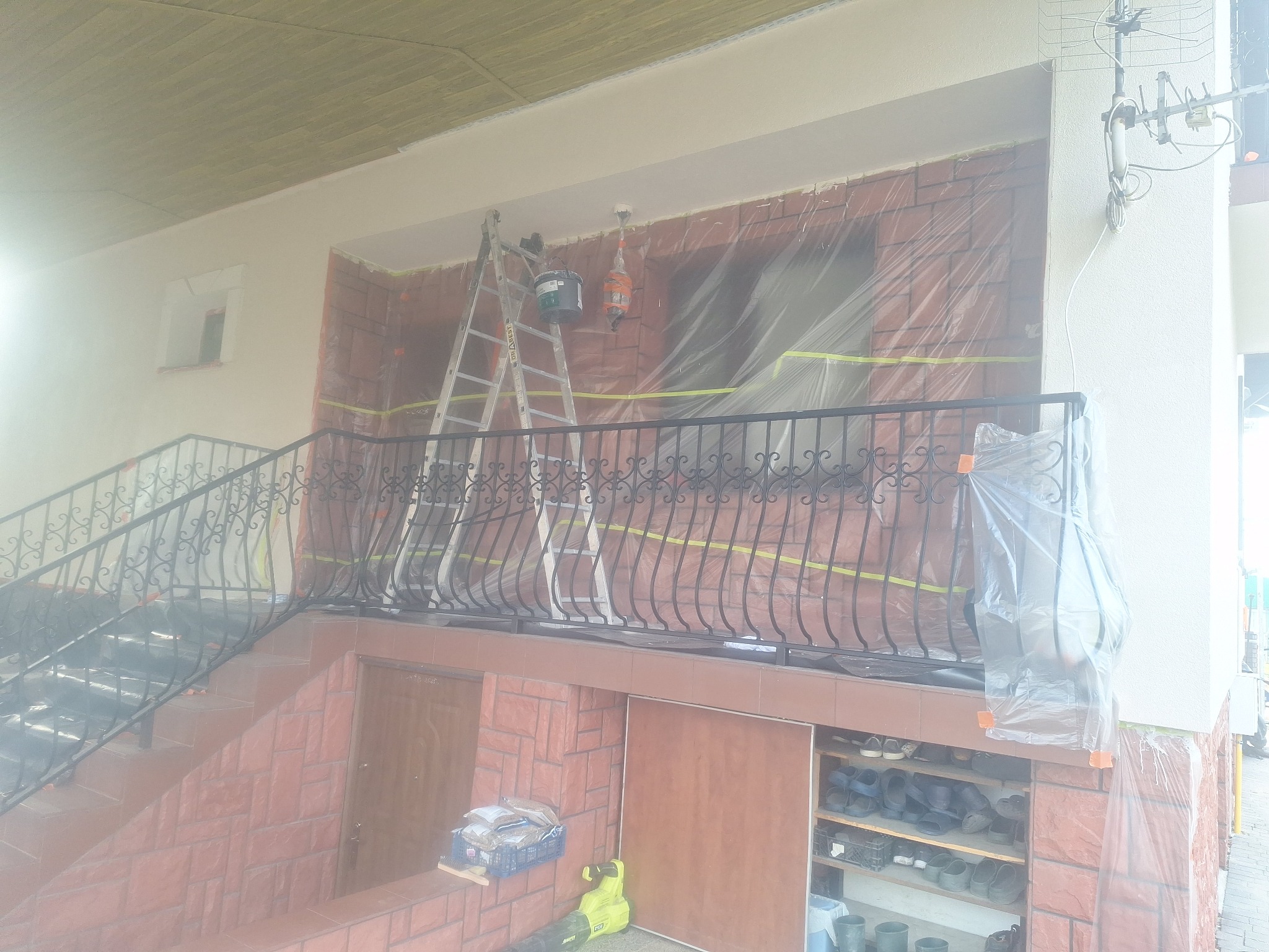 Przygotowania do malowania elewacji domu: zabezpieczona folią ściana z oknami, drabina, wiadro z farbą. Widoczne schody z kutą balustradą i narzędzia.